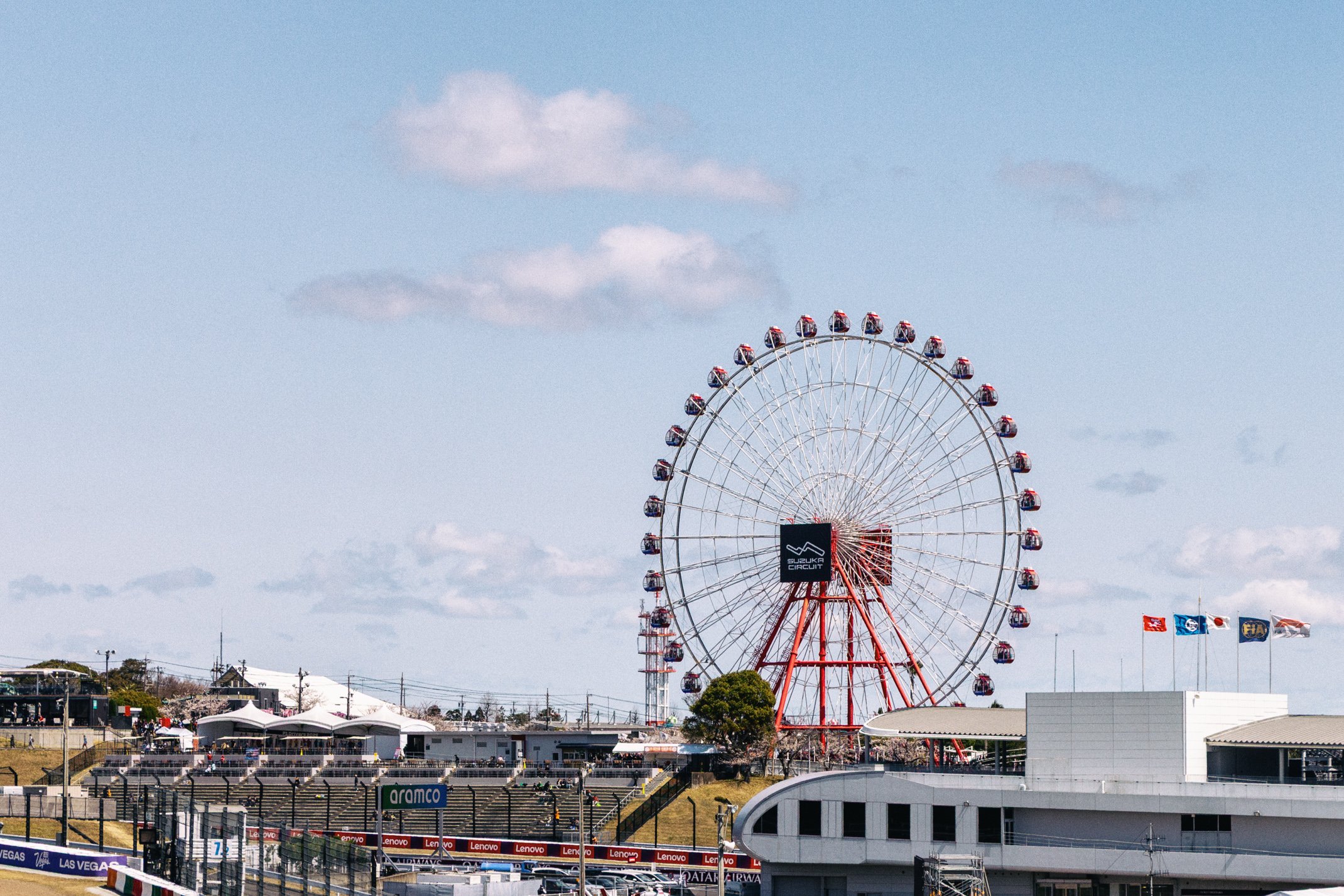 Large red Ferris wheel at a fairground with blue sky and scattered clouds, flags, and buildings nearby.
