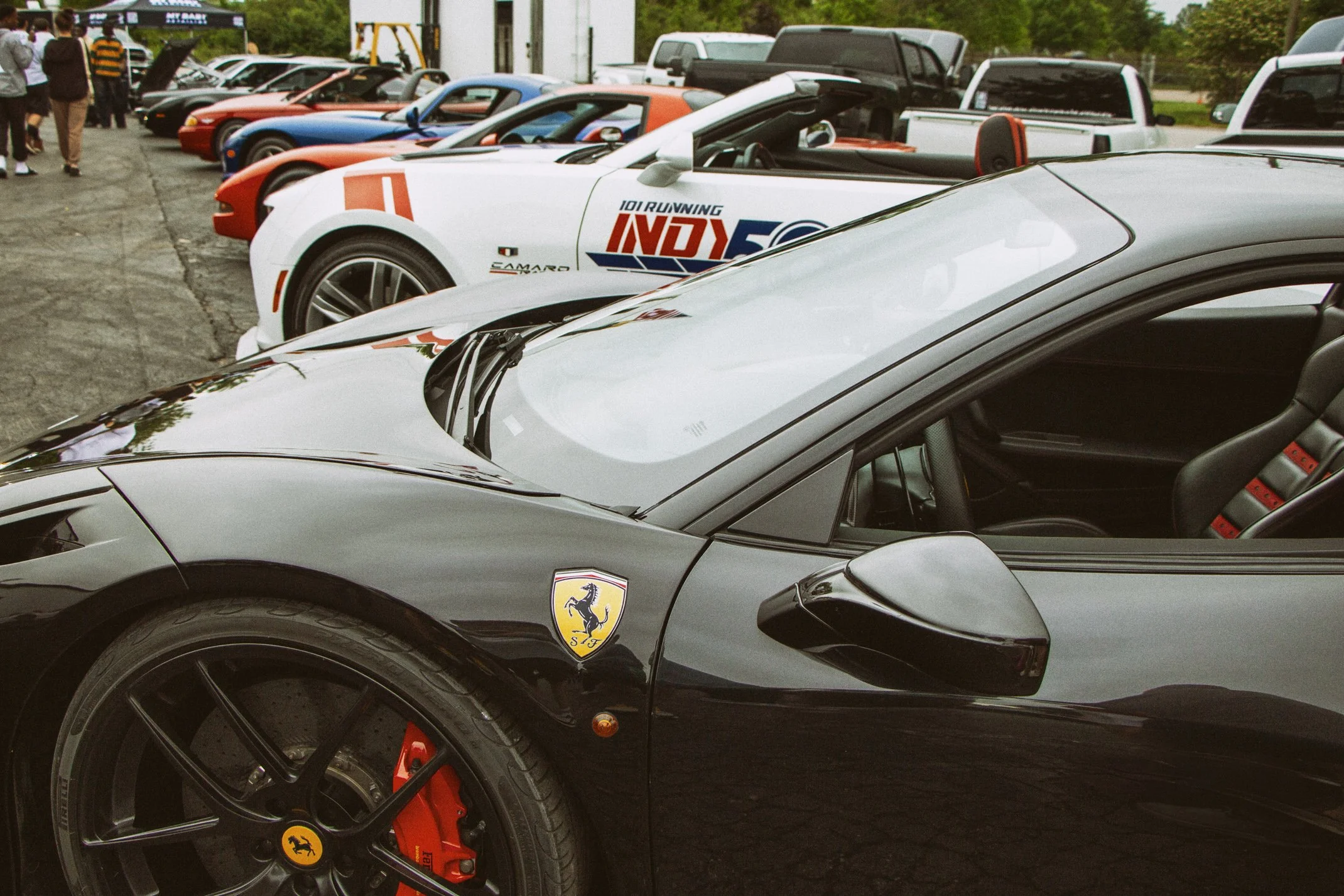 Row of luxury sports cars, including a black Ferrari with a Ferrari logo on the wheel, a white car with Indy 500 graphics, and several other sports cars parked outdoors with people walking nearby.