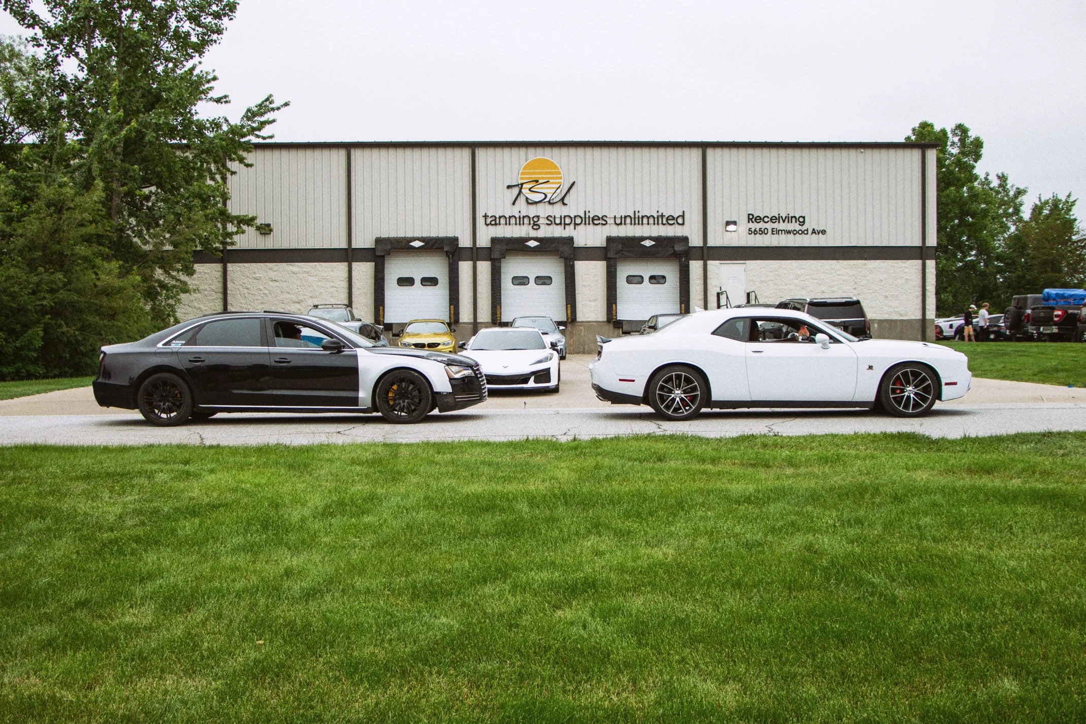 A building with the sign TSQ Tanning Supplies Unlimited, three garage doors, and a parking lot with several cars, including a gray and white sports car in the foreground. There are trees on both sides of the building and a grassy area in front.
