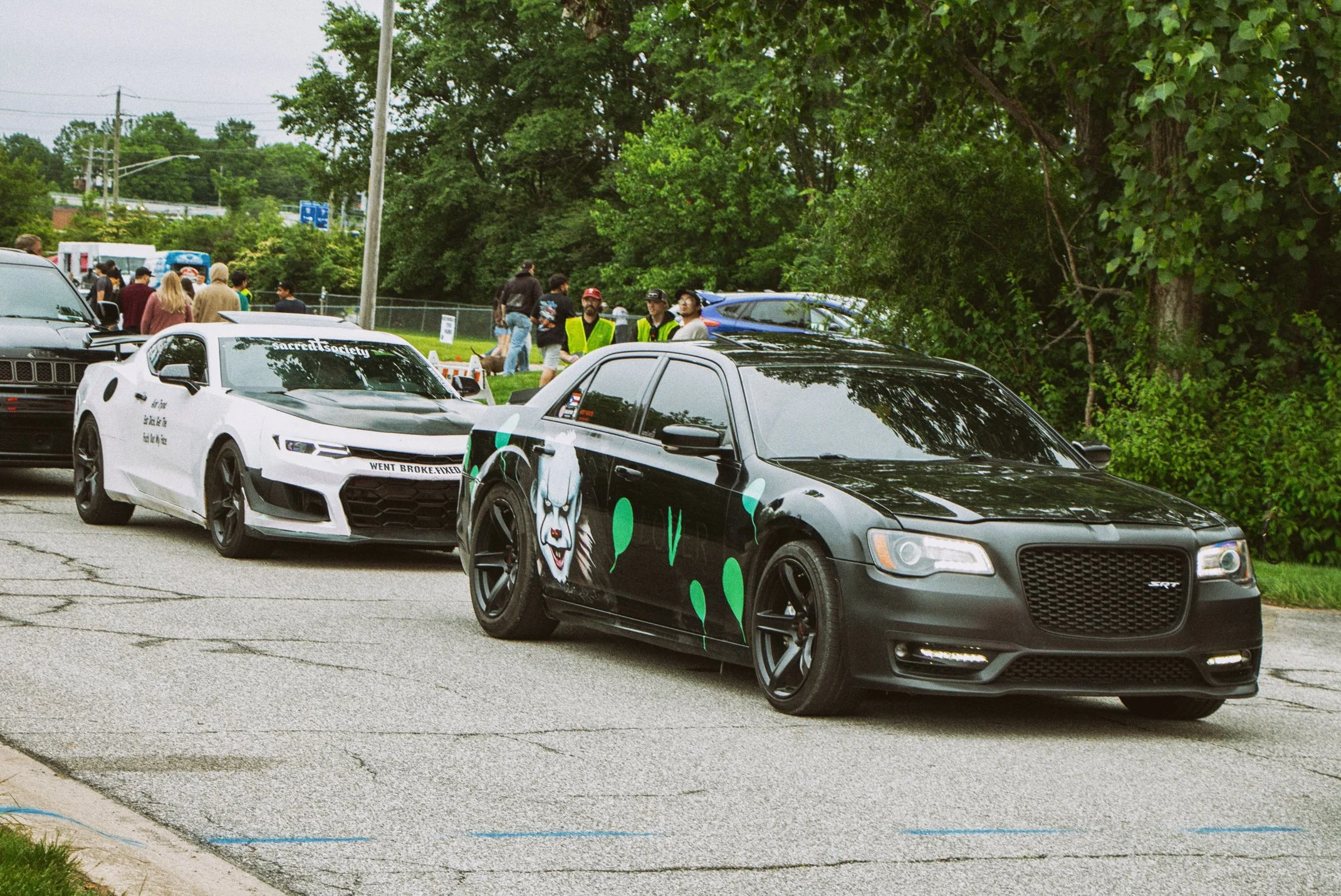Several cars parked outdoors with a group of people standing nearby, some wearing high-visibility vests, under green trees on an overcast day.