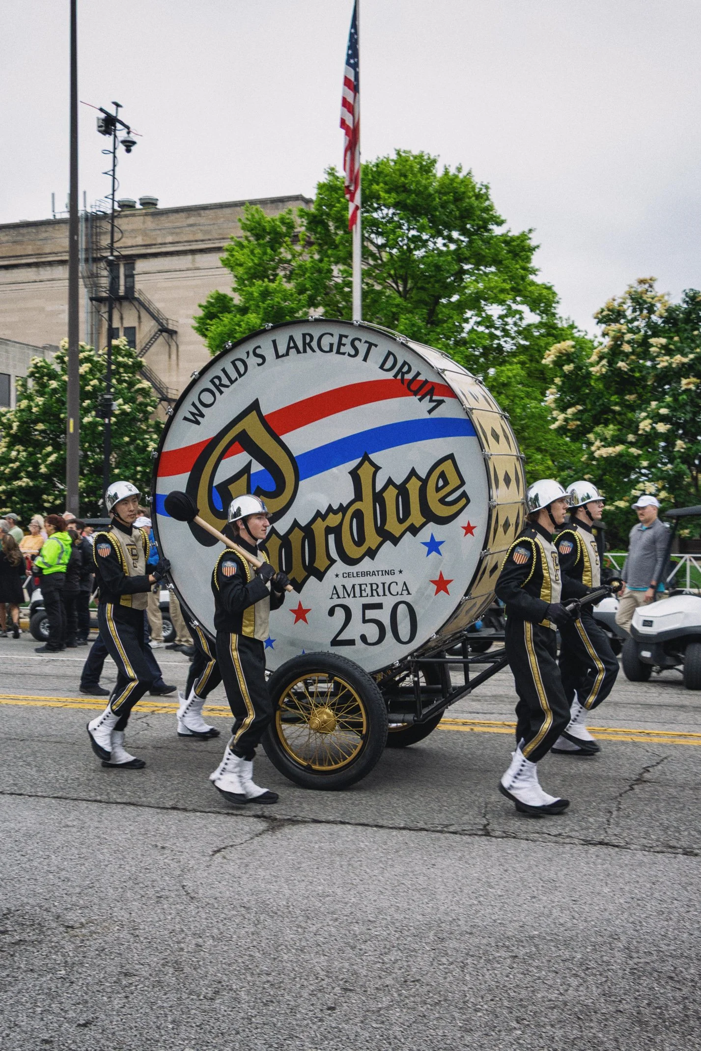 A parade float celebrating the 250th anniversary of America features a large drum with the words "World's Largest Drum" and "Celebrating America 250". Uniformed drumline members are marching alongside the float on a city street, with spectators and g
