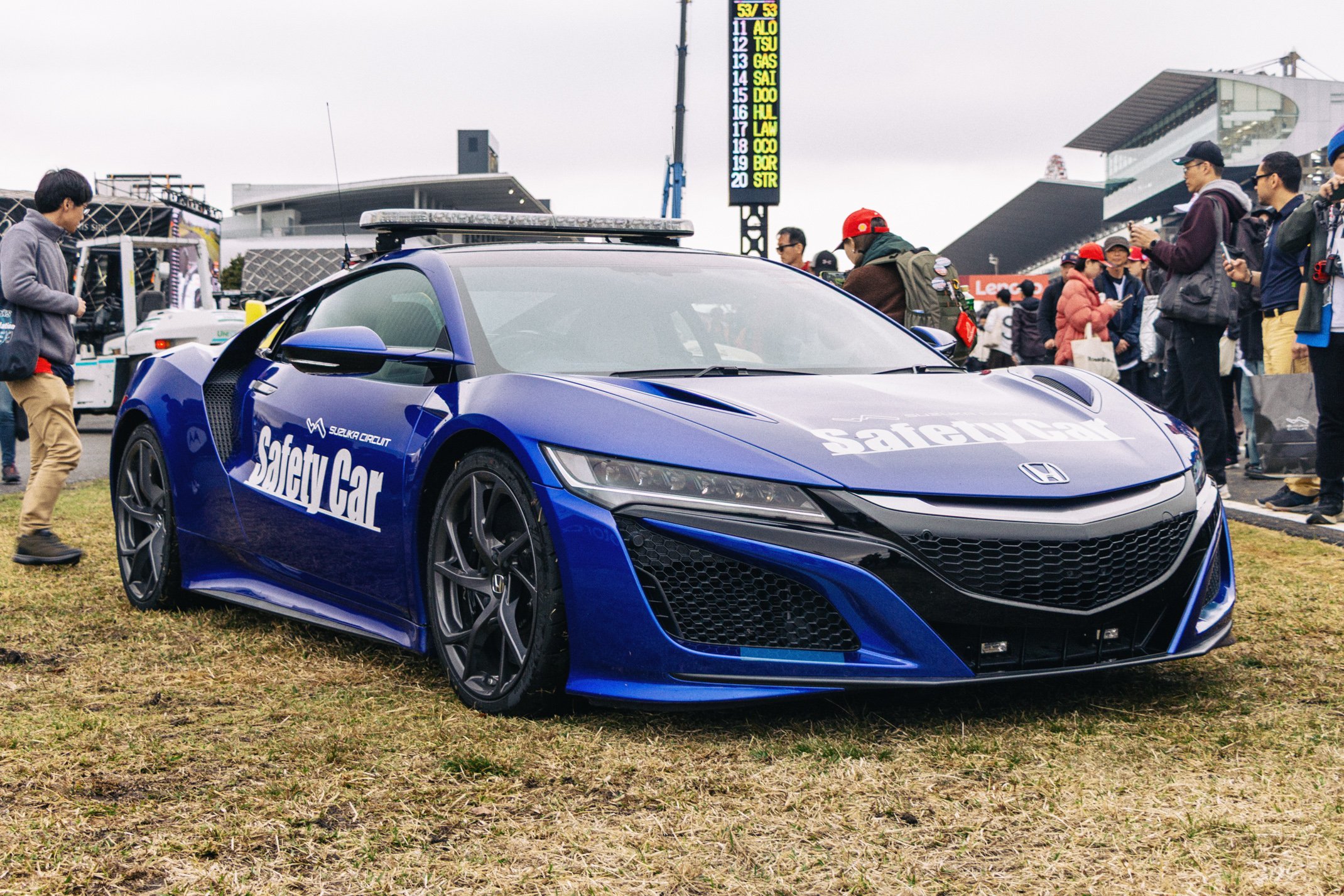 Blue Honda safety car with the words "Safety Car" written on the side, parked on grass at a motorsport event with spectators and a digital scoreboard in the background.