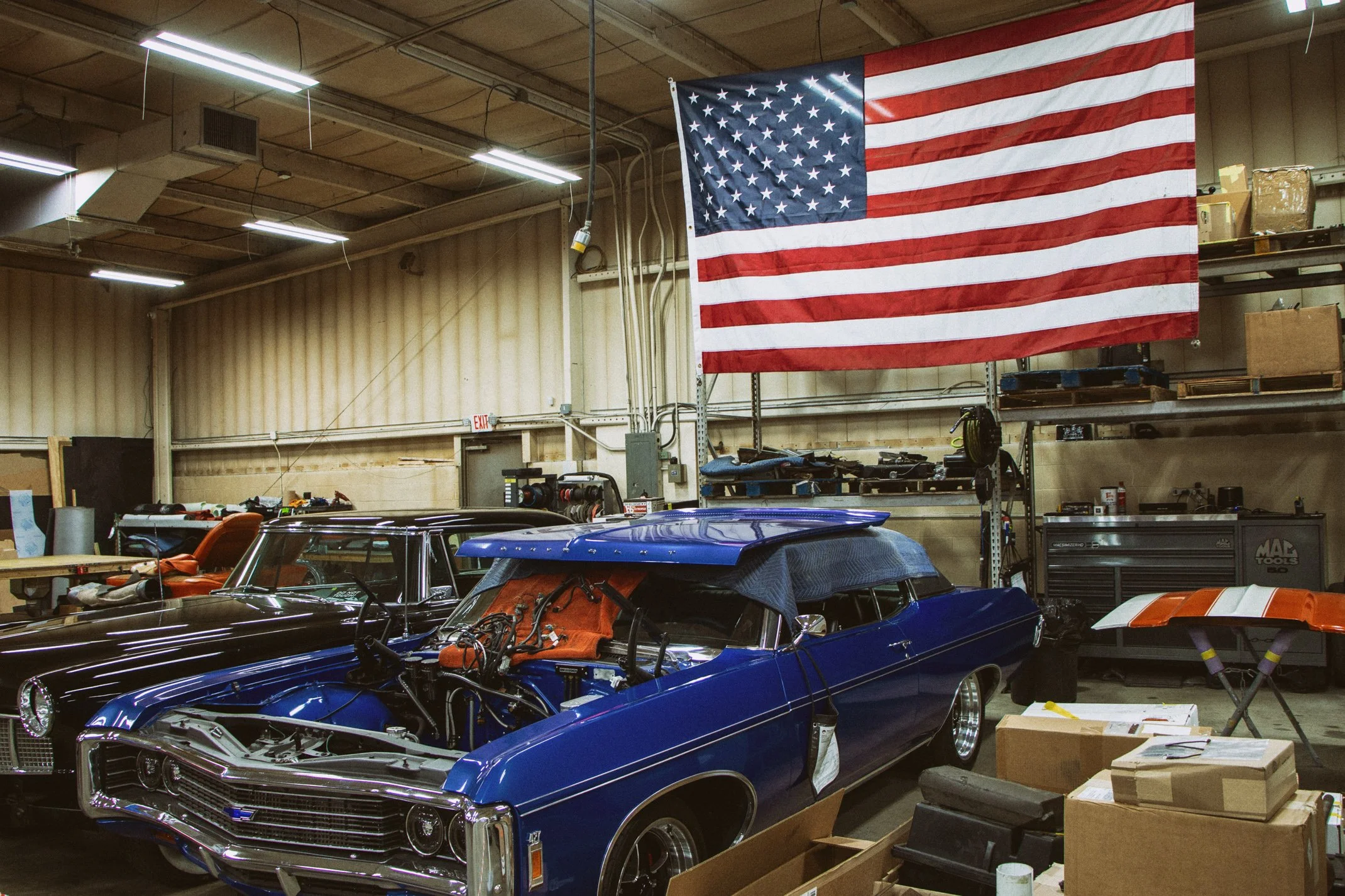 An indoor garage or workshop with a large American flag hanging on the wall. Several classic cars are parked inside, with tools and equipment around. The ceiling has exposed pipes and fluorescent lights.