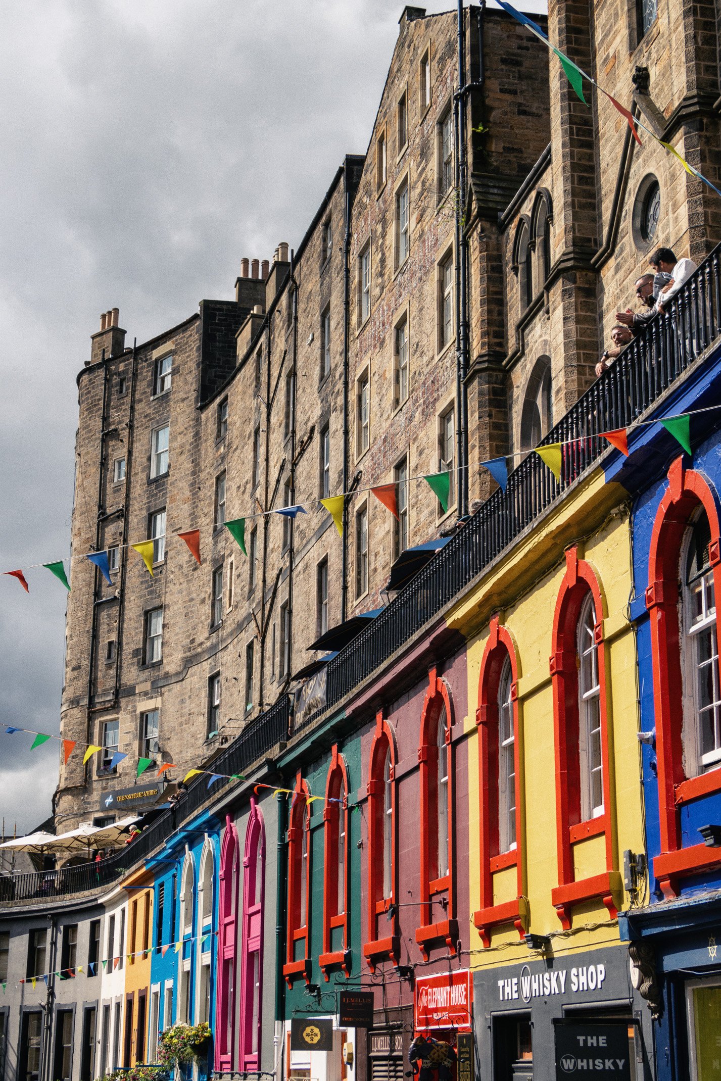 Colorful buildings with red, yellow, pink, and blue window frames along a street, with multi-story brick buildings above and decorative bunting hanging across the street.