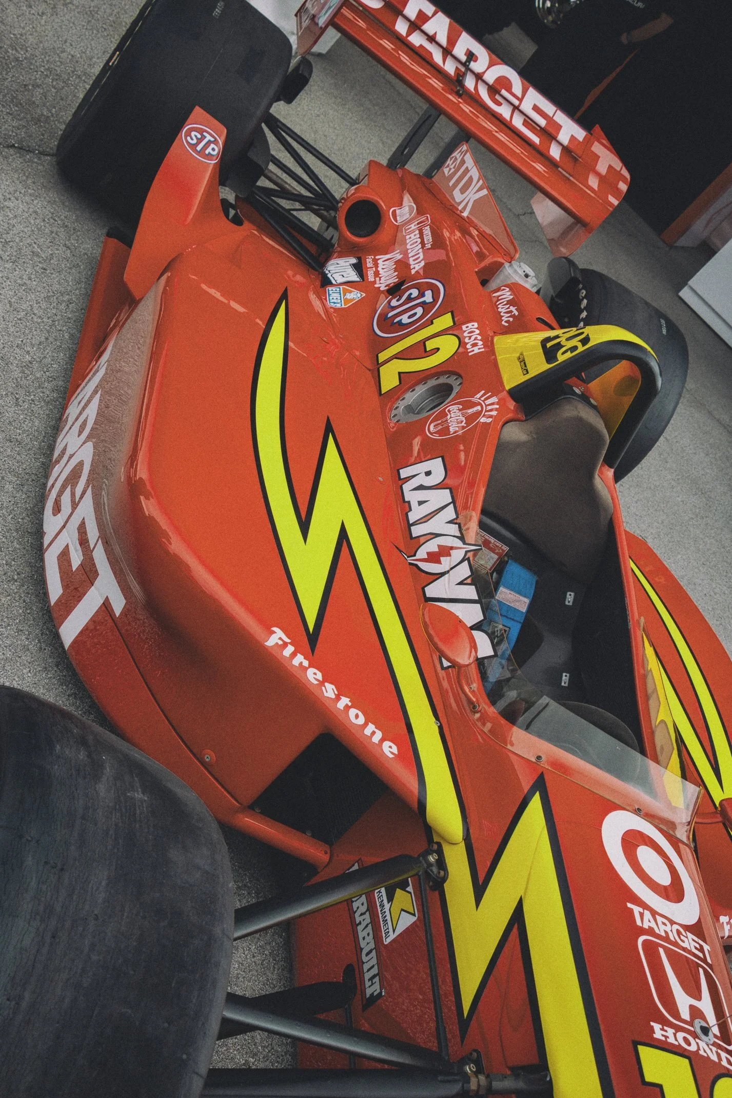 A bright red open-wheel race car with sponsorship logos, large rear tires, and a single-seat cockpit, positioned on a garage floor.