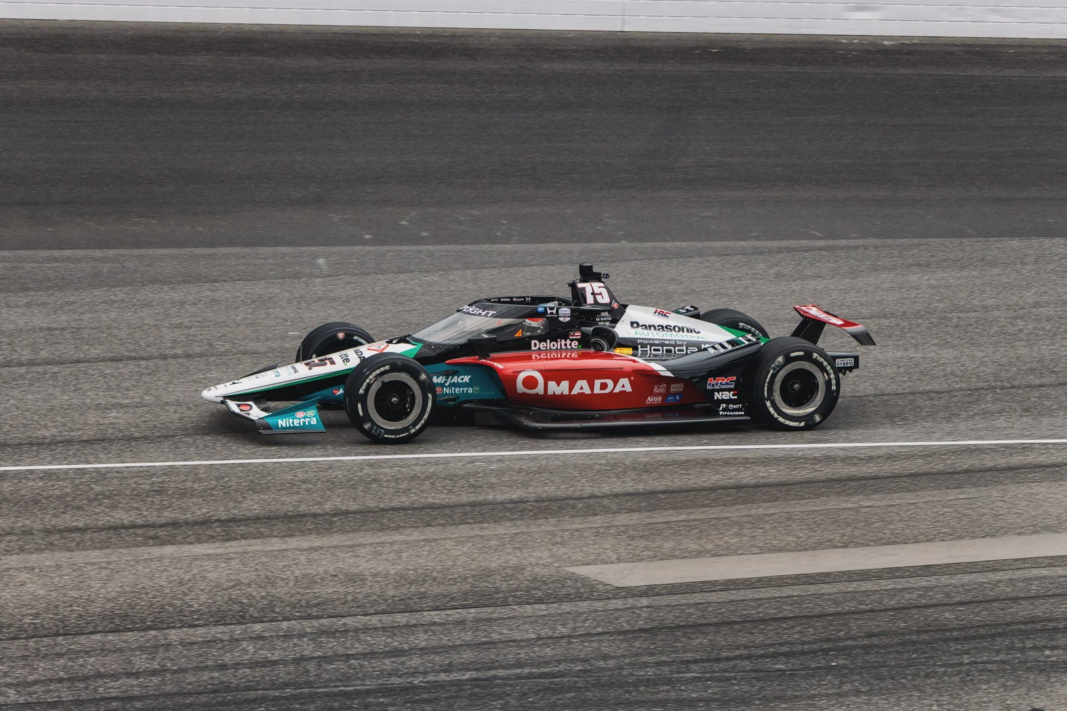A race car on a race track with sponsor logos, including Daimler, Panasonic, and Honda, moving at high speed.
