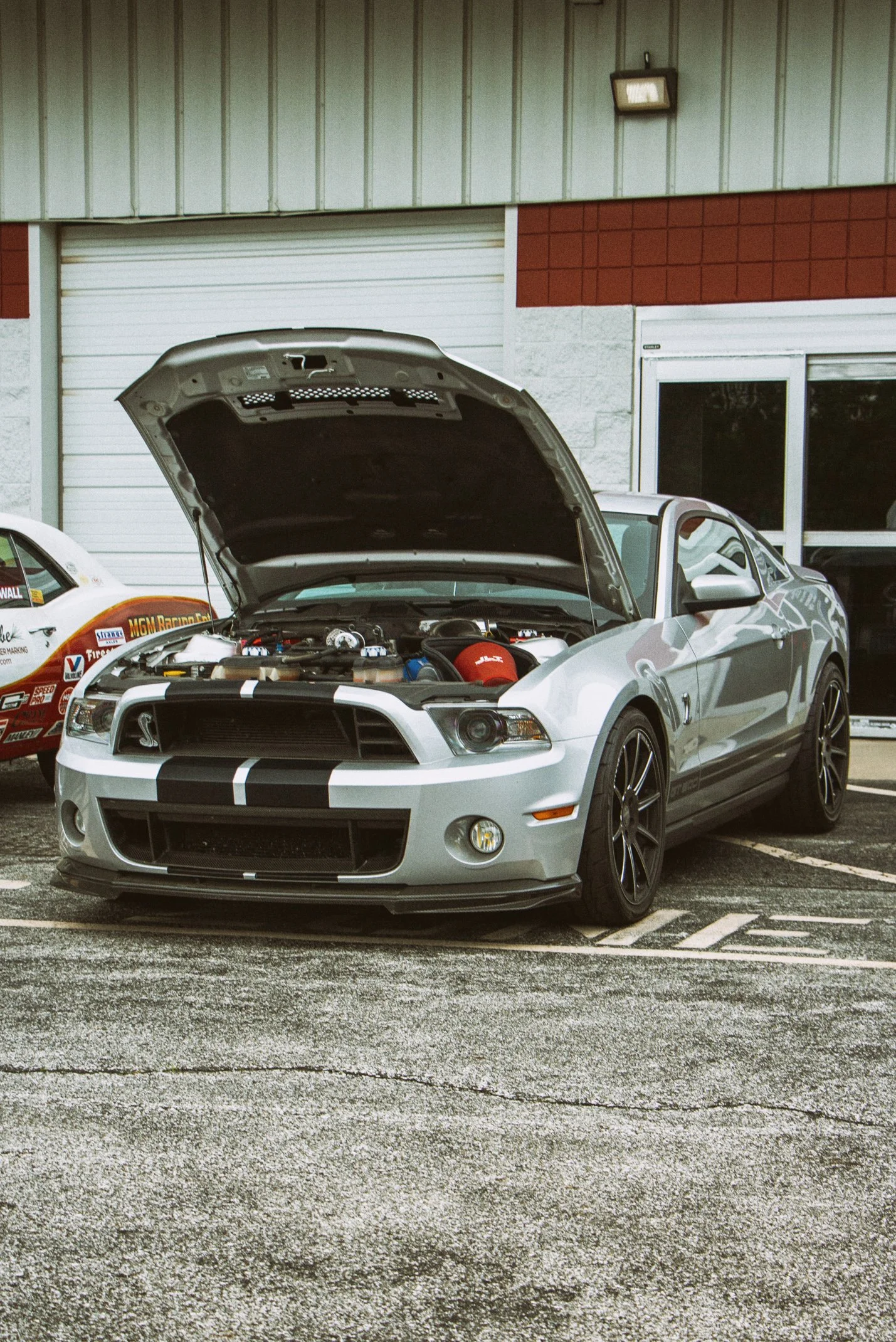 A silver Ford Mustang with black racing stripes has its hood open, parked in a parking lot next to a racing car and a building with a red and beige exterior.