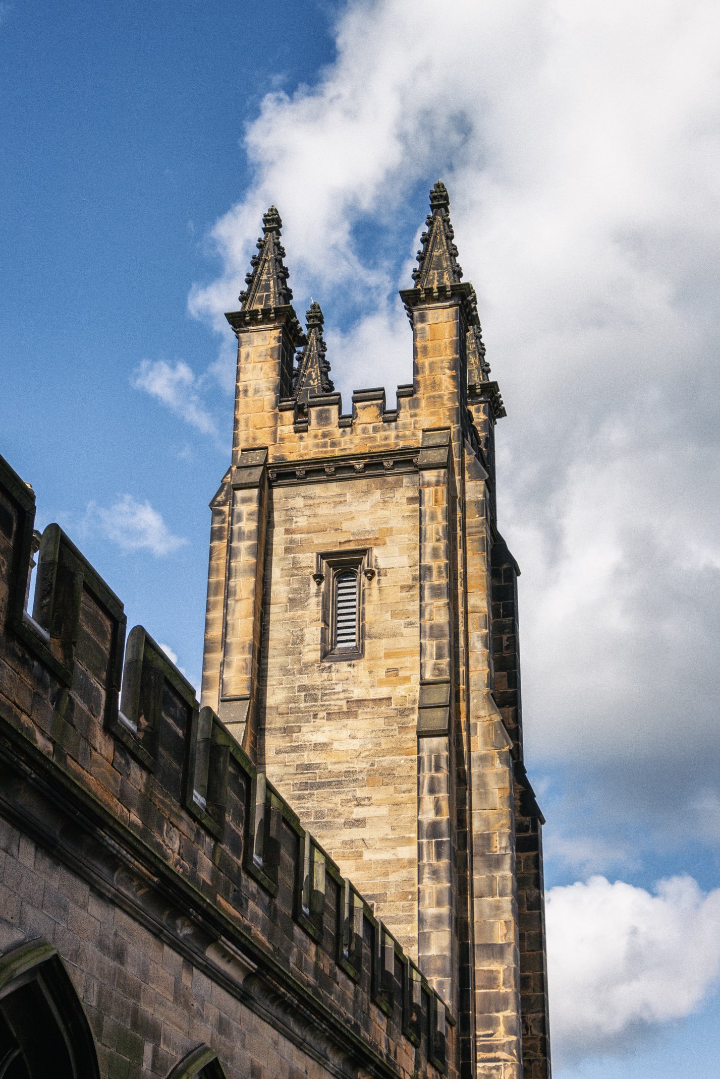 A tall, gothic-style church steeple made of dark stone with two pointed spires and ornate details, set against a partly cloudy sky.