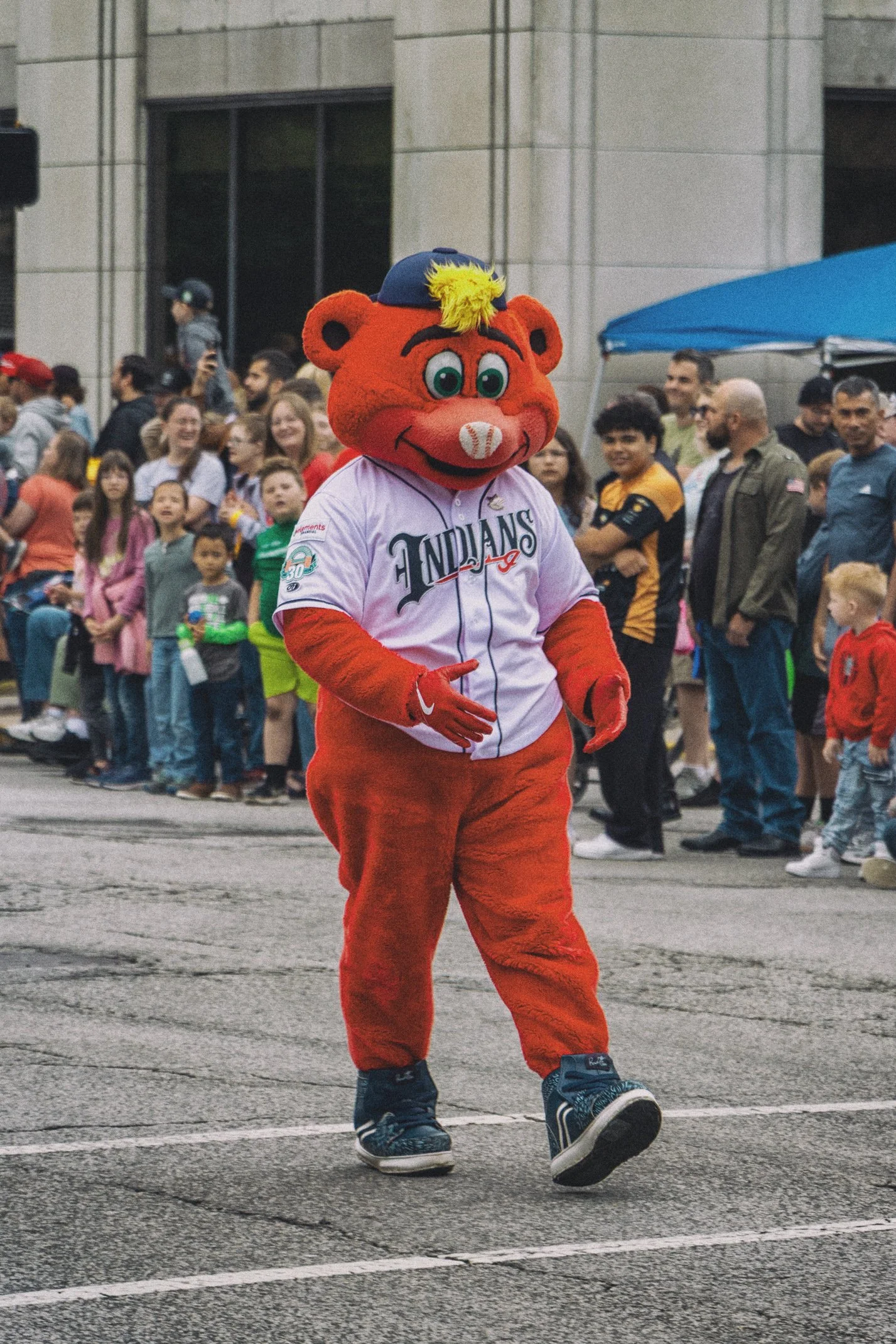 A mascot dressed as a red bear wearing a baseball jersey and a cap with a yellow tuft, walking on a street with a crowd of people watching the parade.