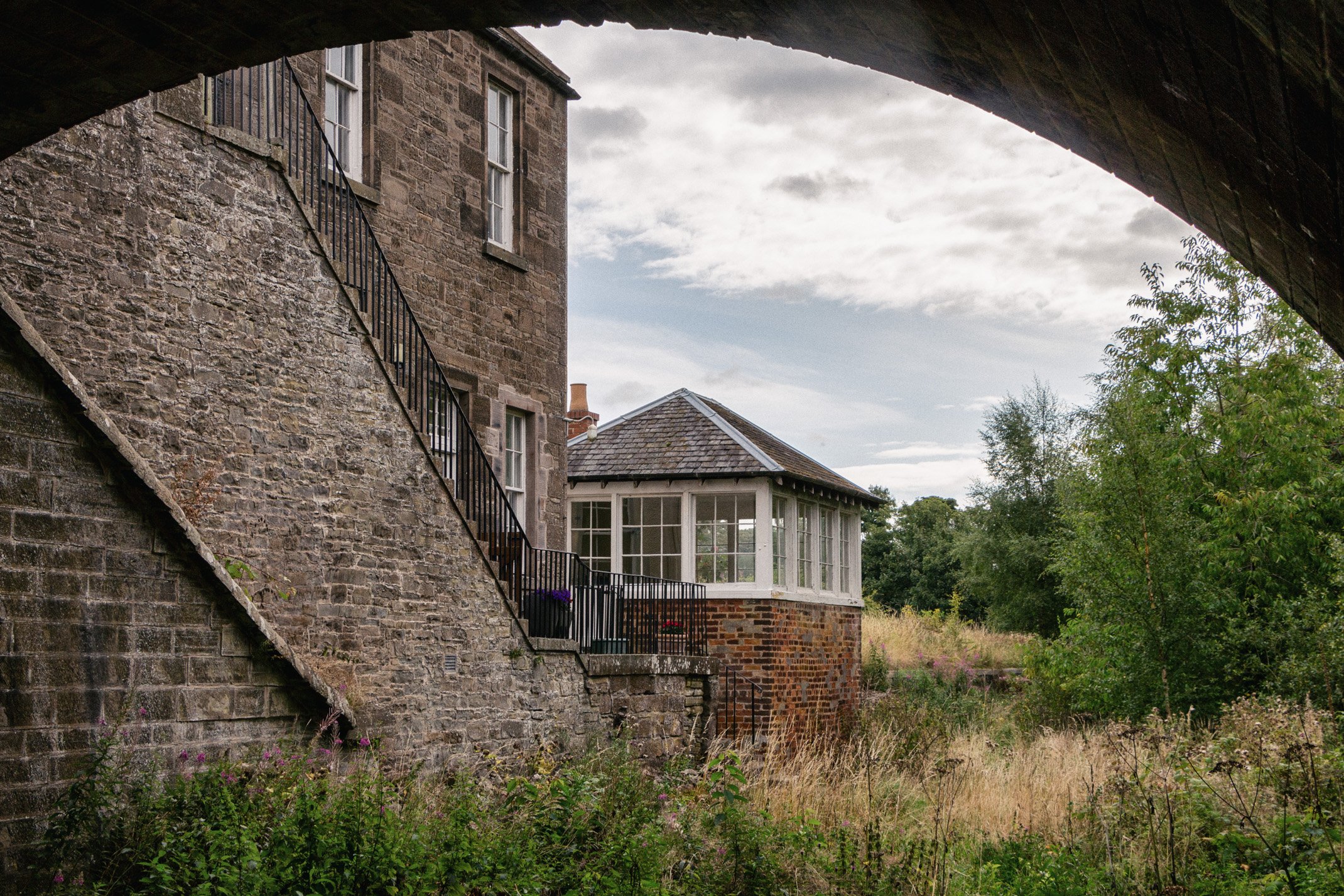 View of a stone and brick house with a staircase, a bay window, and a grassy yard, seen from underneath a bridge.