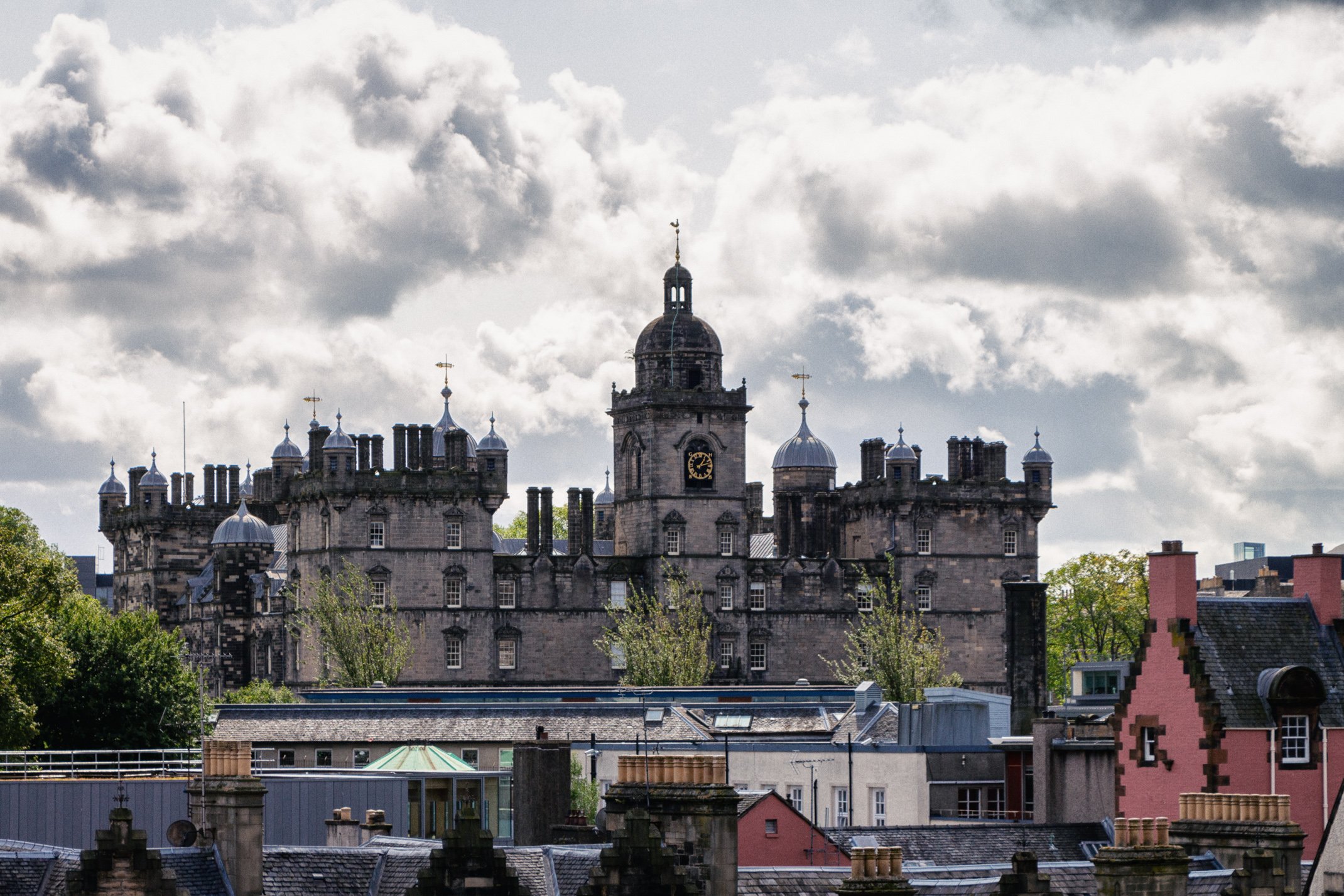 A historic castle with multiple towers and a clock tower, set against a cloudy sky. The foreground features rooftops of surrounding buildings and trees.