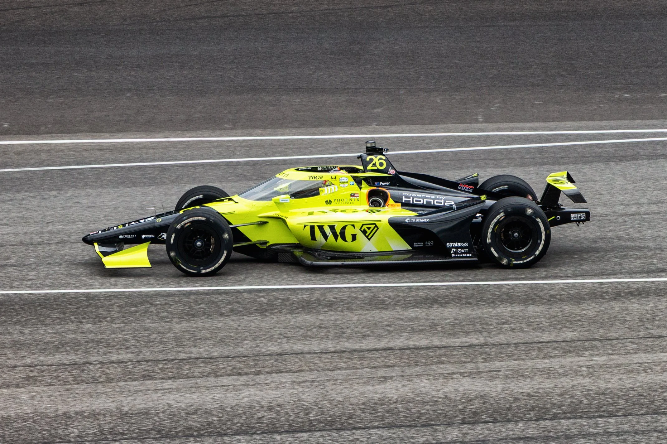 A green and black IndyCar race car with the number 26 driving on a racetrack.