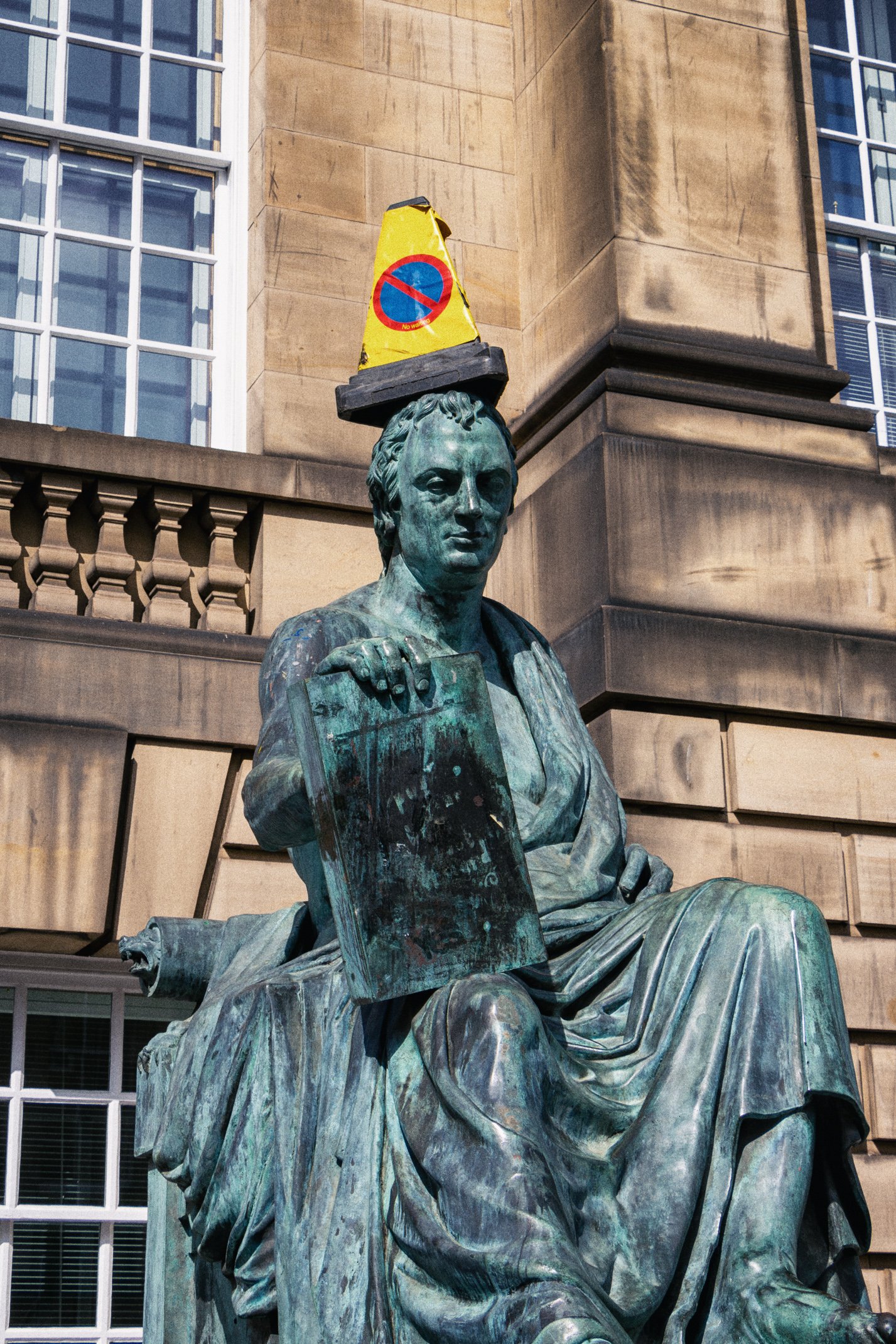 Bronze statue of a seated woman holding a tablet, with a construction cone on her head that has the no parking sign. The background shows a building's brick and window facade.