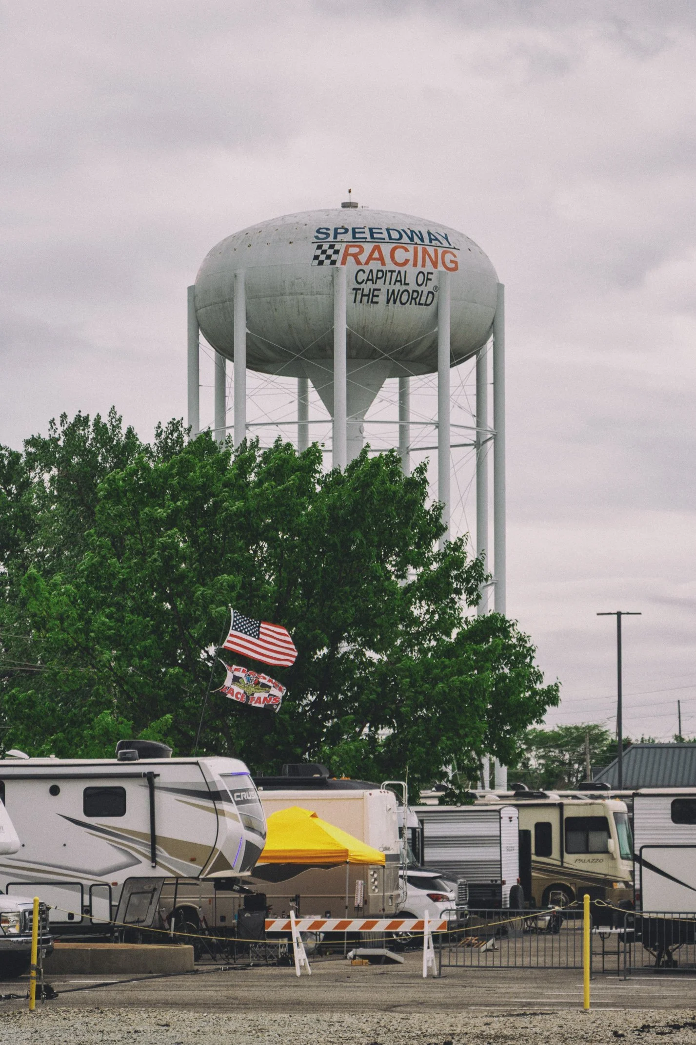 A large white water tower with the words 'Speedway Racing Capital of the World' painted on it, standing against a grey, overcast sky. In the foreground, there are RVs, trailers, and tents in a parking lot, along with some American flags and flags for