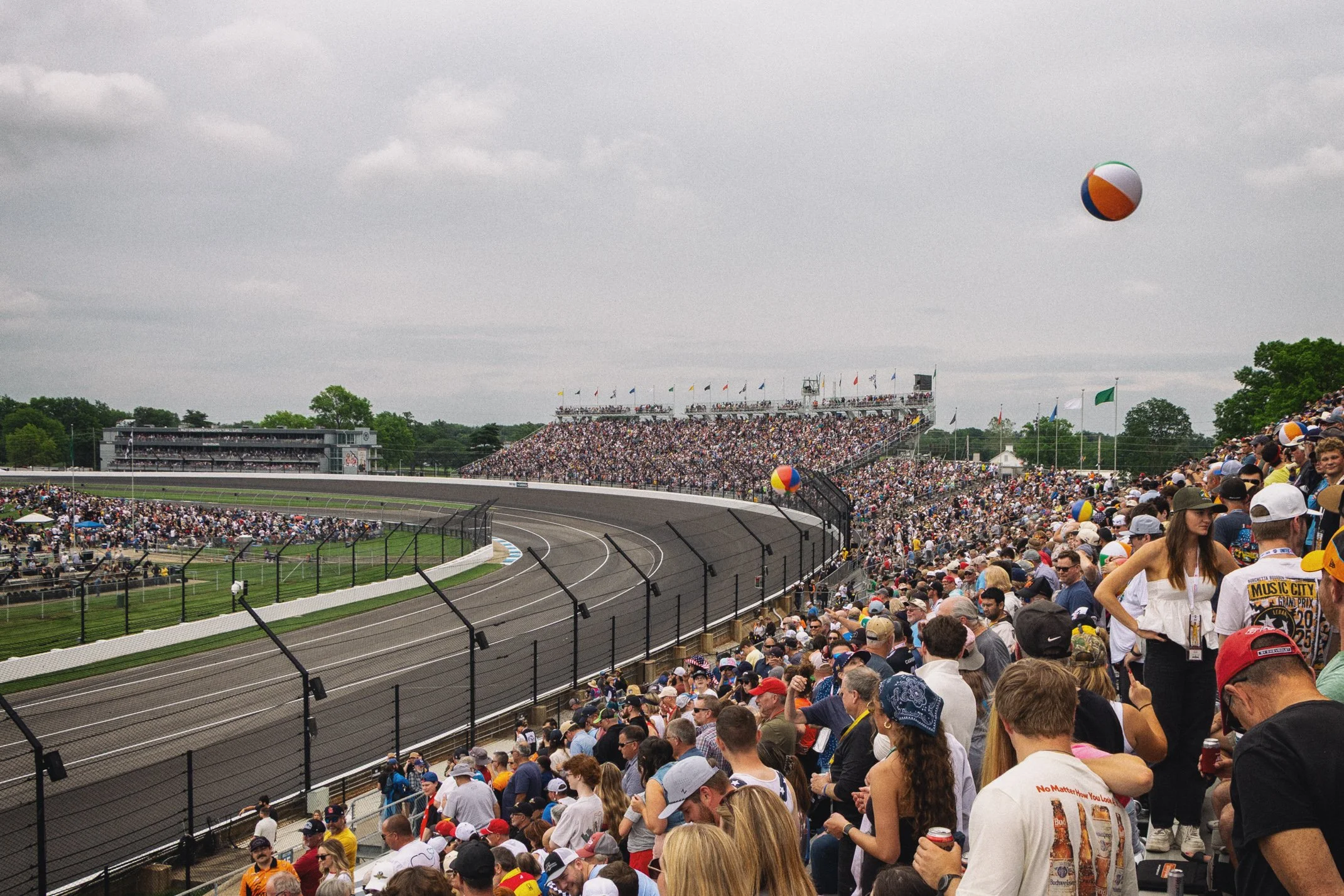 View of a crowded outdoor race track with spectators in the stands and along the fence, a large inflatable ball in the sky, and overcast weather.