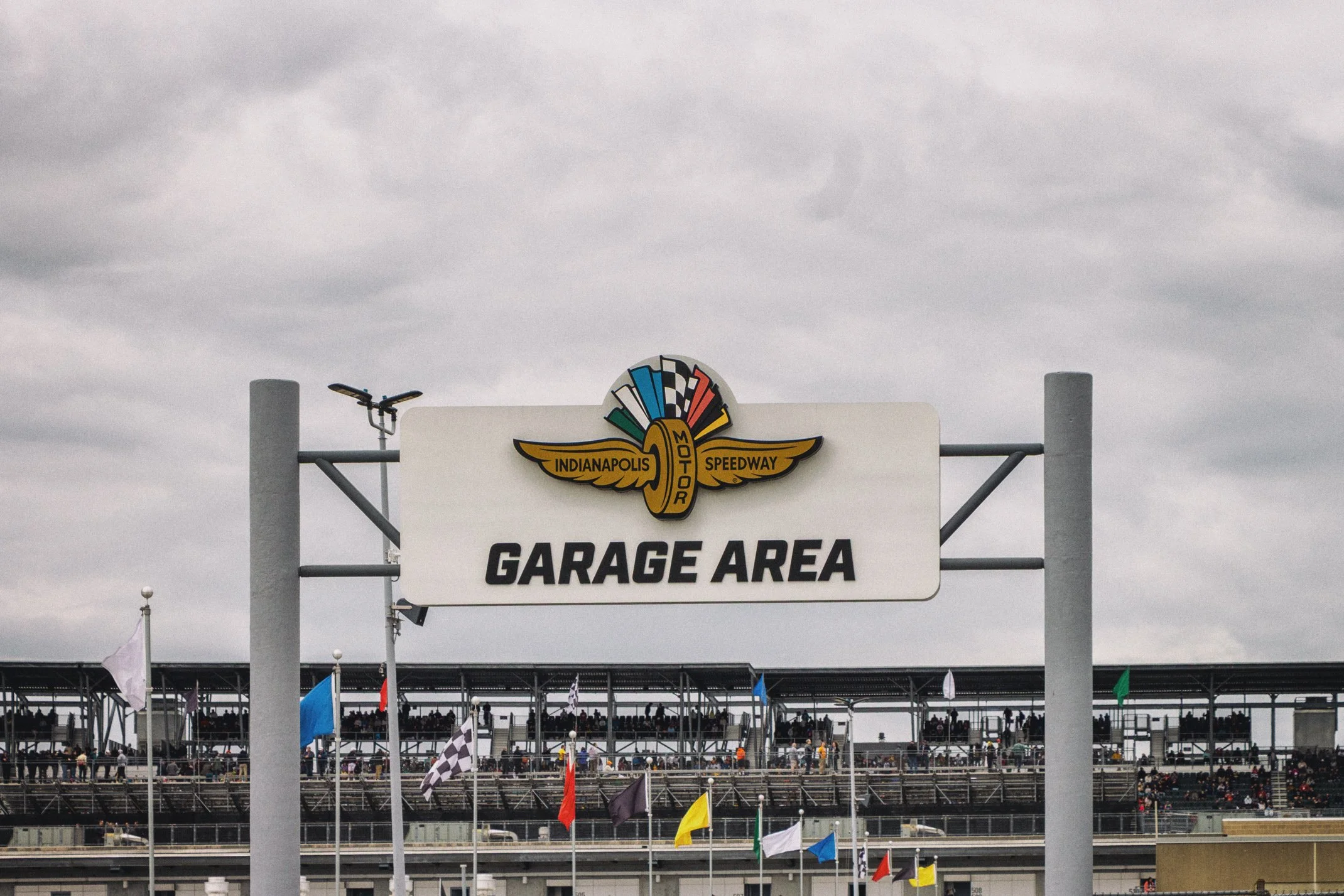 Sign at an Indianapolis Motor Speedway garage area with the Speedway logo and checkered flag design, overcast sky overhead, with crowd and flags visible in the background.