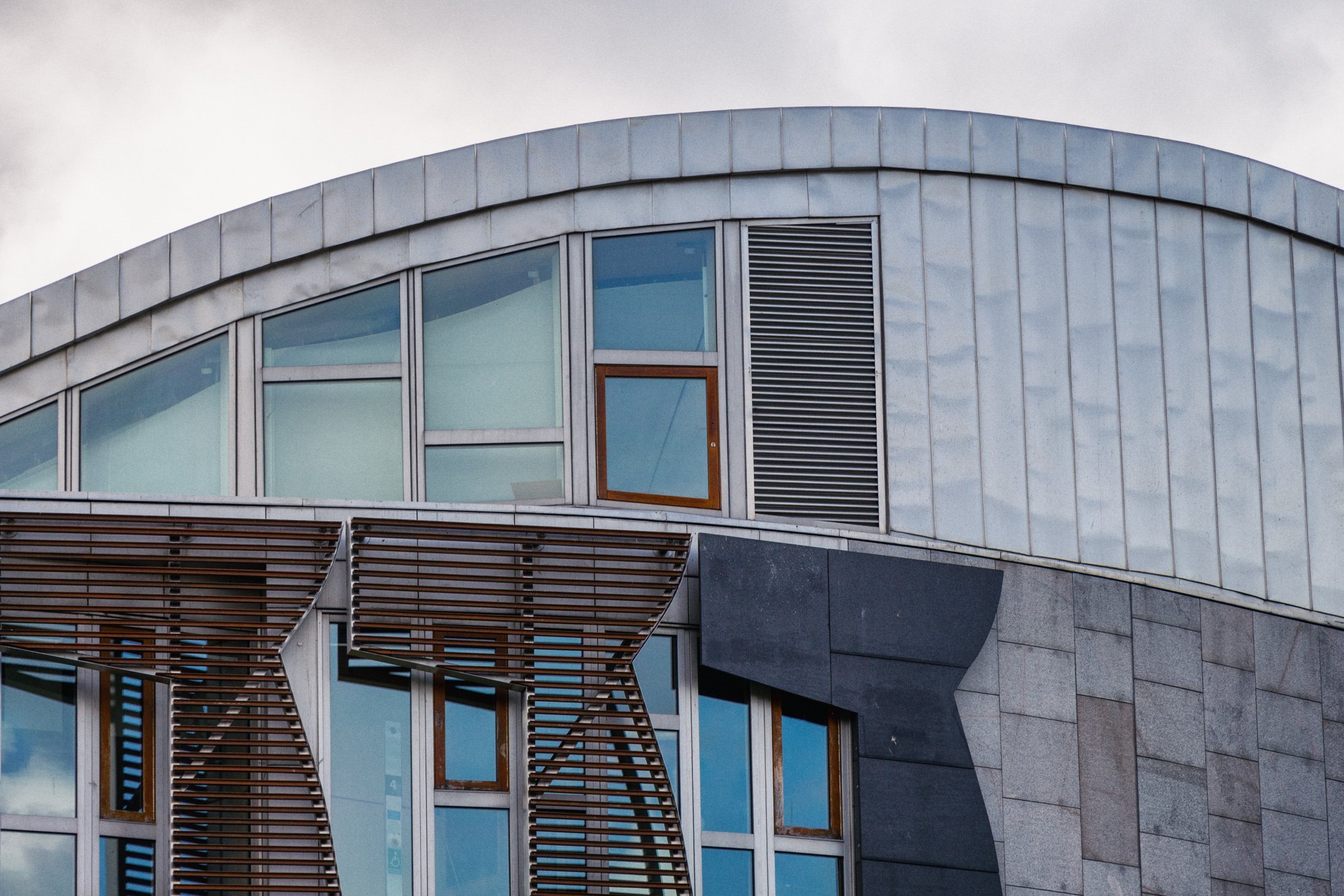 Close-up of a modern building with curved glass windows, wood and metal accents, and an overcast sky background.