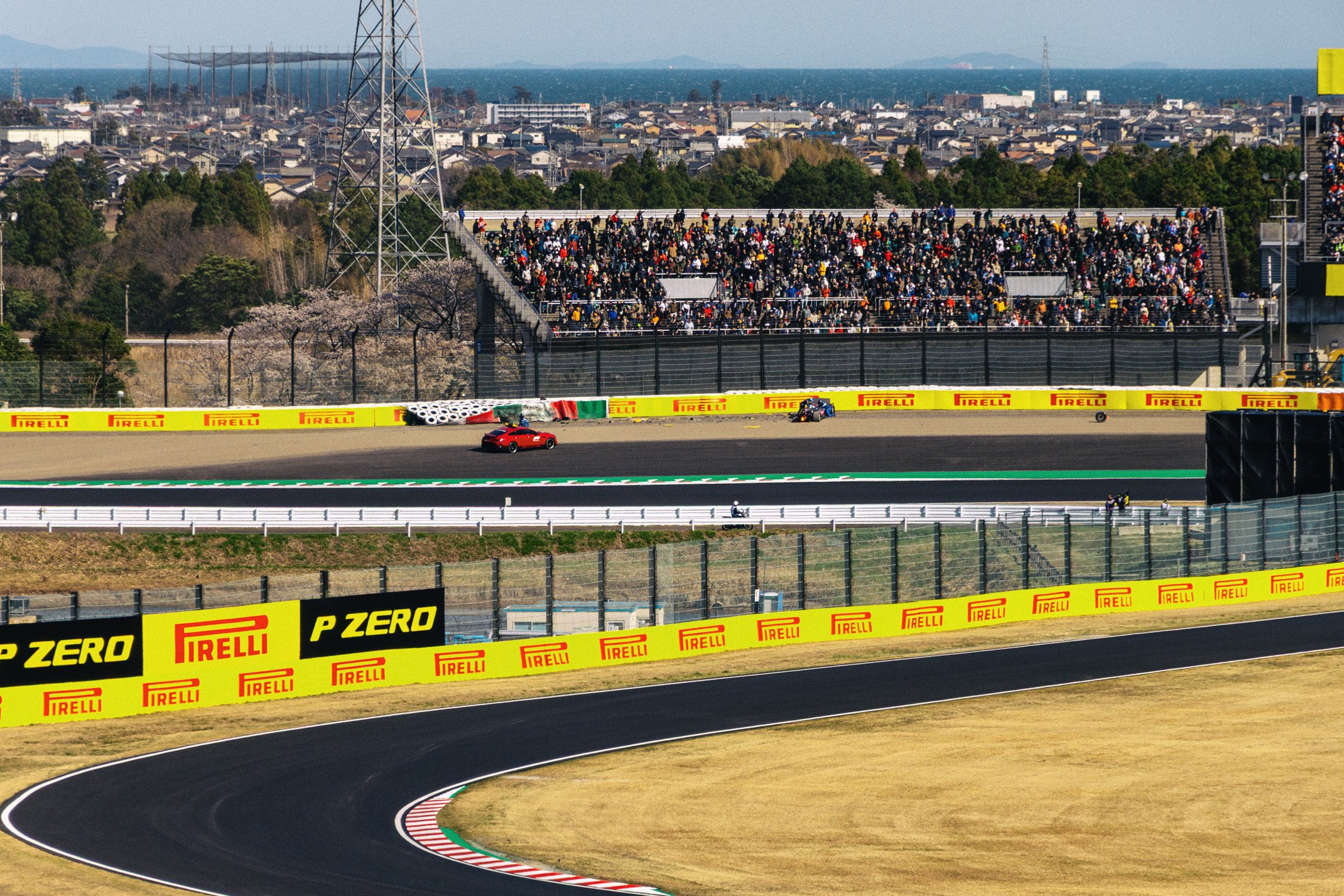 A race track with a red car and a black racing vehicle passing by. Large crowd in the grandstands, and advertising banners for Pirelli on the fence and trackside. The background shows a suburban landscape with houses, trees, and a distant ocean.