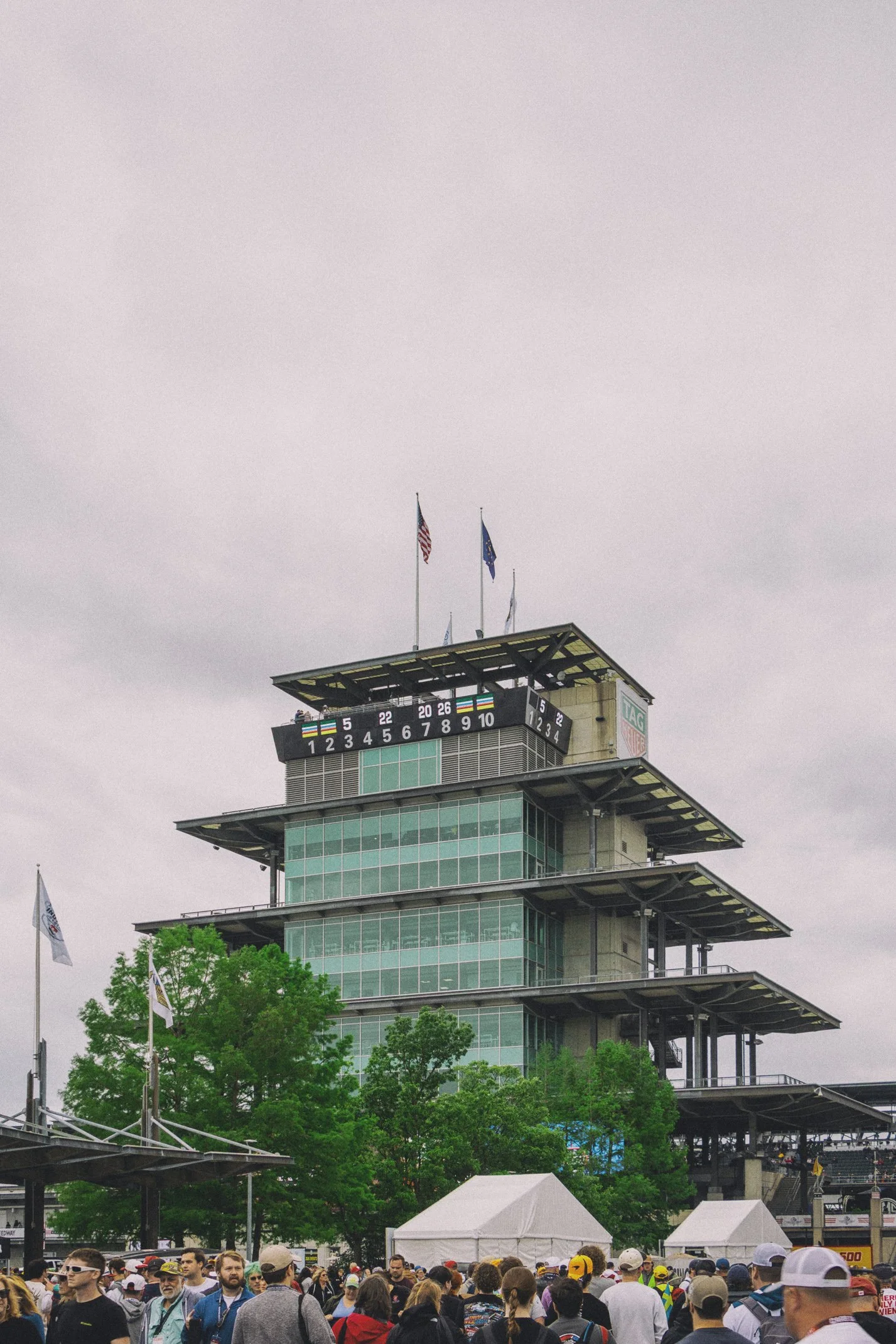 Crowd of people outside at a public event with a stadium tower in the background, topped with flags and a digital scoreboard.