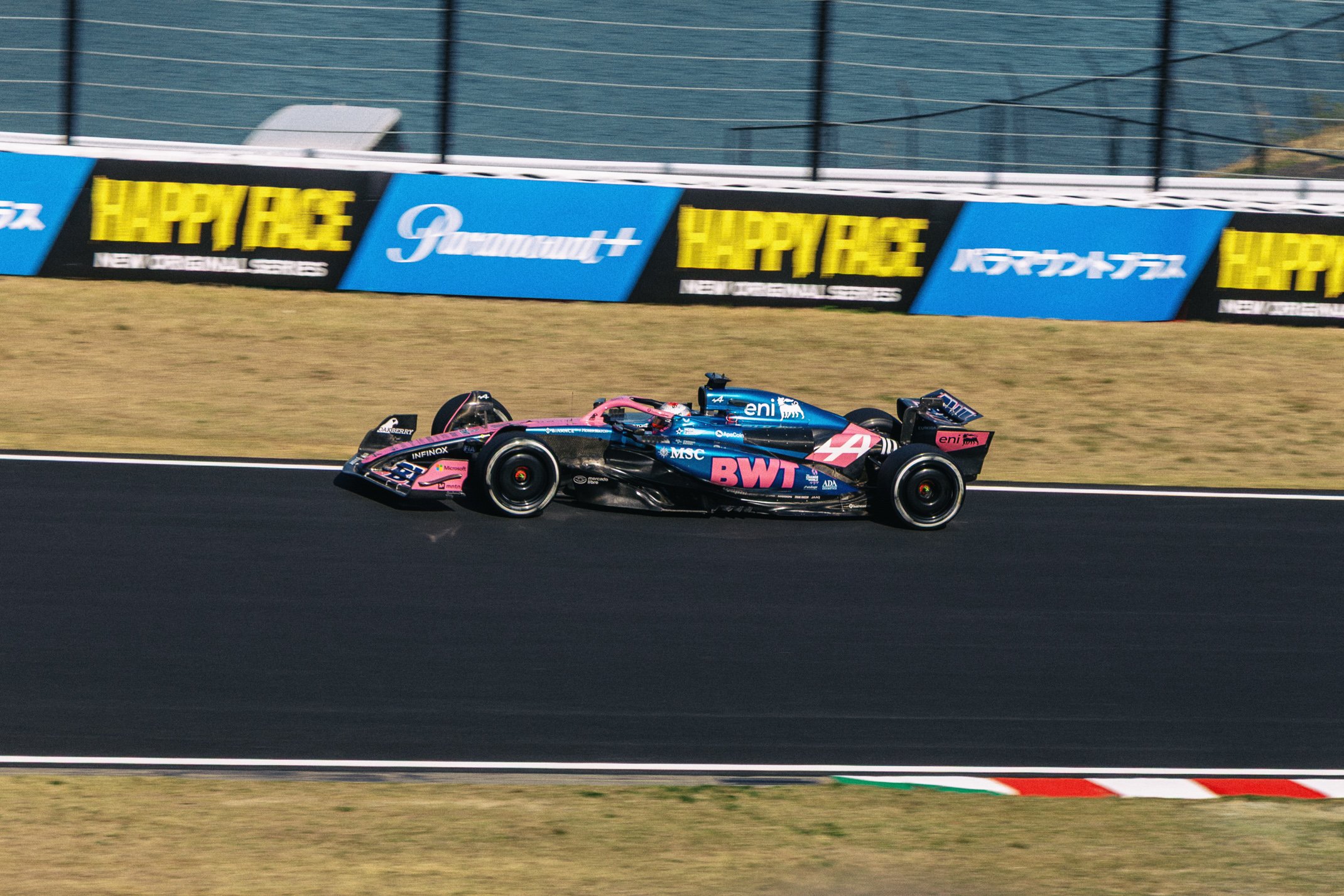 A blue and pink Formula 1 race car on a racetrack with advertising banners in the background.