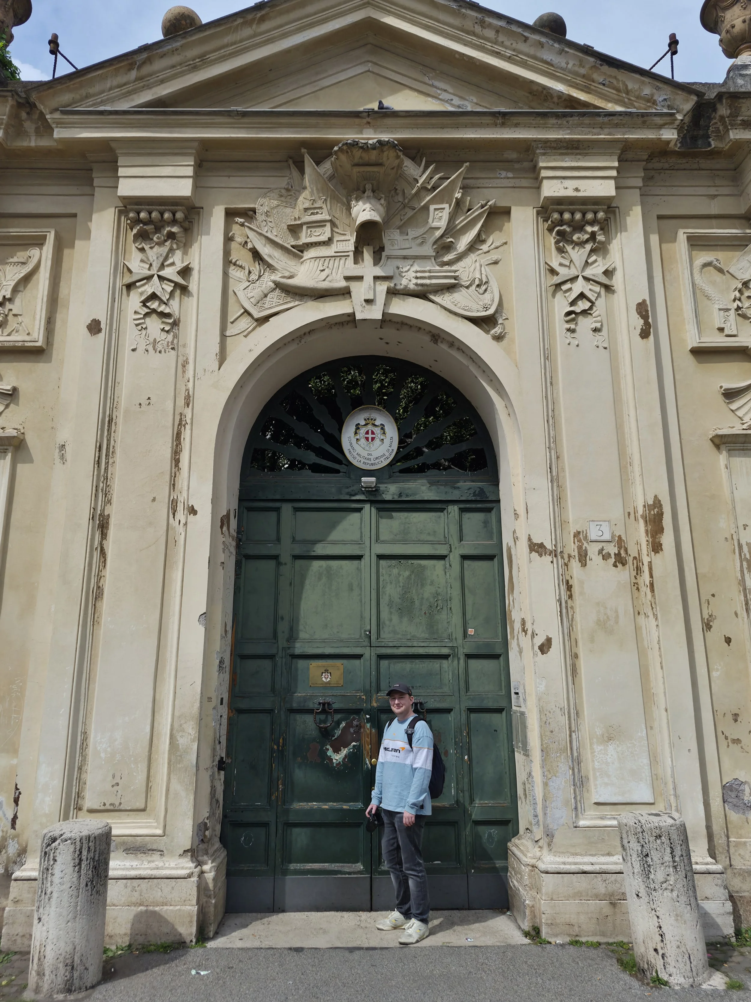 A man standing in front of an old, large, green wooden door with ornate stone architecture around it. The architecture features carved details, flags, and a crest with a cross, indicating a historical or governmental building. The building appears weathered and aged.