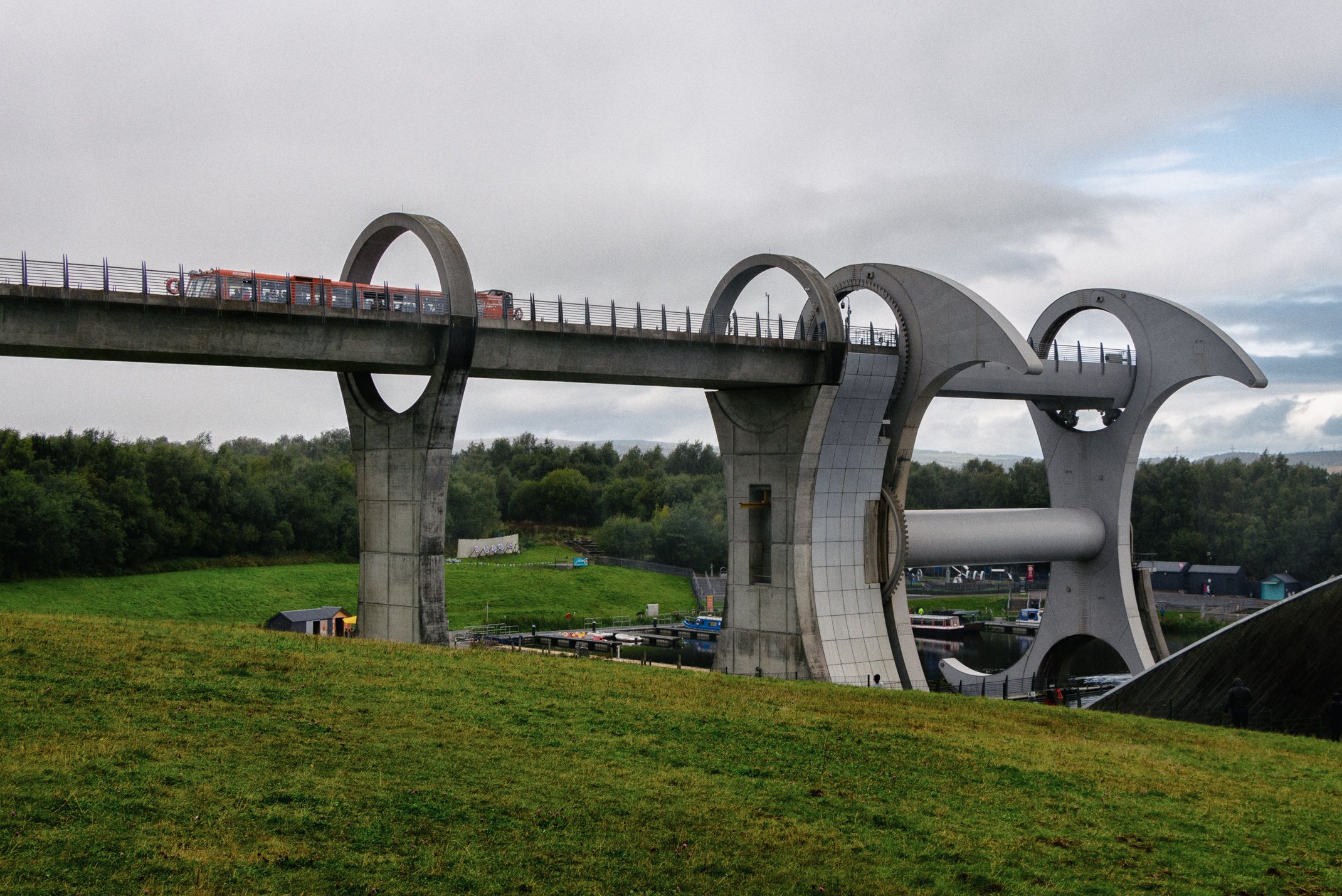 The Falkirk Wheel, a large rotating boat lift, located outdoors with grassy land in the foreground and trees in the background. The structure has a unique design resembling a giant wheel with two circular arms and a connecting canal.