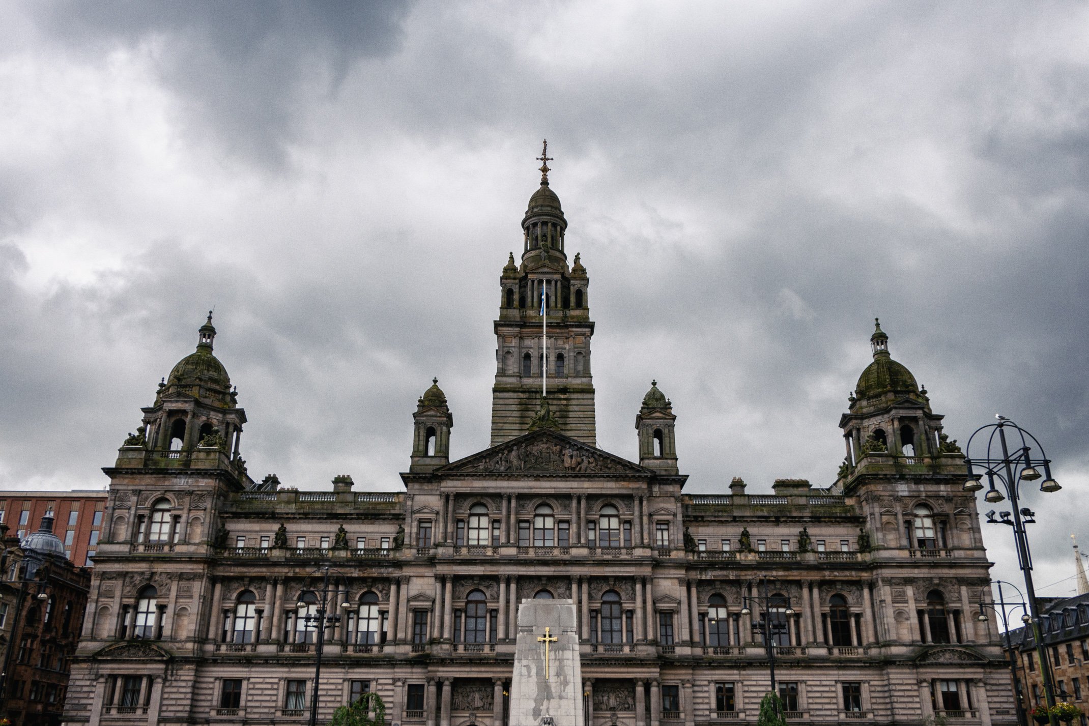 A large historic government building with multiple towers and domes, under a cloudy sky, featuring a cross monument in front.