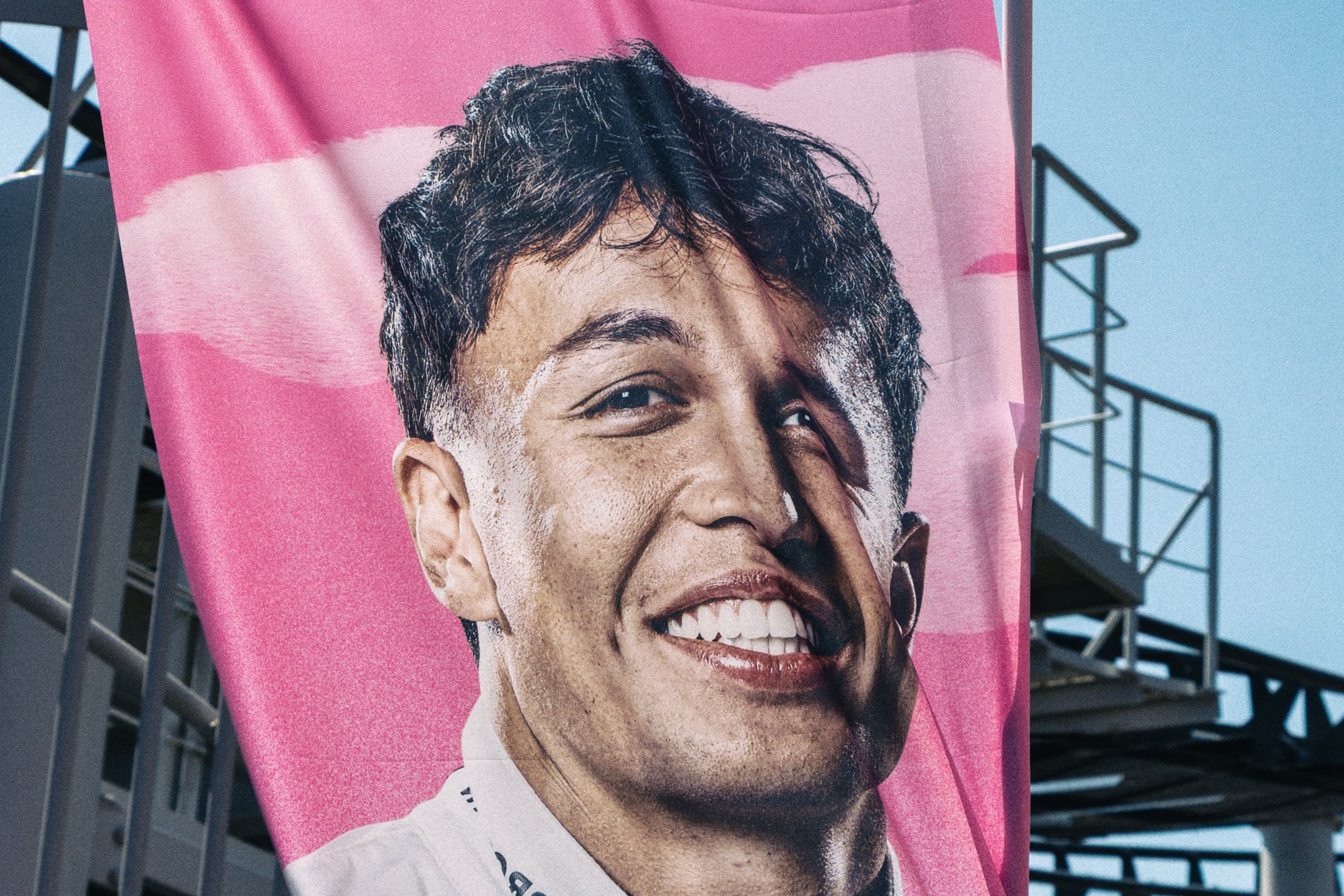 A large billboard with a close-up image of a smiling woman with short dark curly hair, wearing a white top, against a pink background with white clouds, outdoors with a metal structure and blue sky in the background.