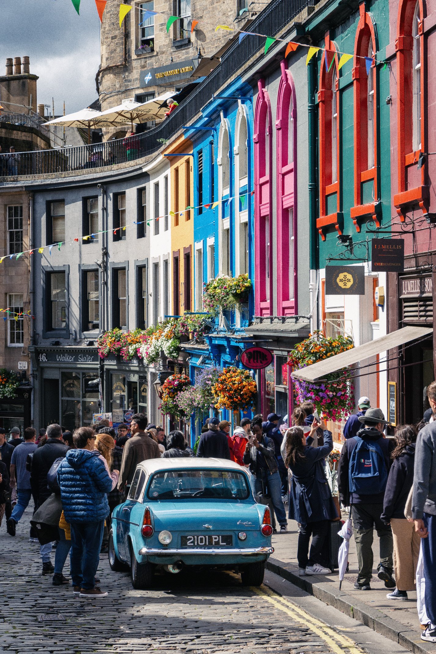 Colorful row of buildings with pink, blue, yellow, and orange facades on a busy street filled with people, a vintage blue car, and floral decorations hanging outside shops.