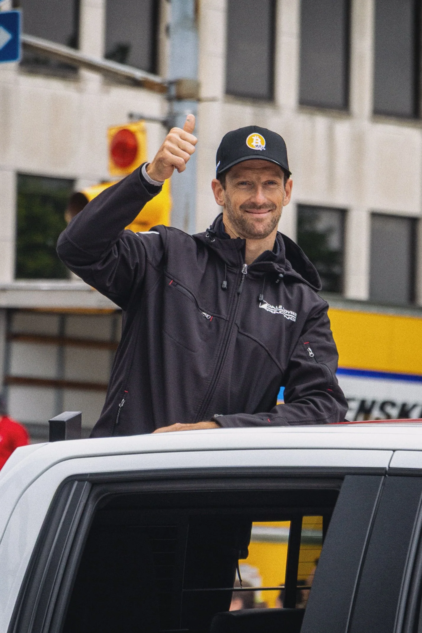 A man in a black jacket and cap with a Bitcoin logo is giving a thumbs up while standing on top of a white vehicle during a race event.