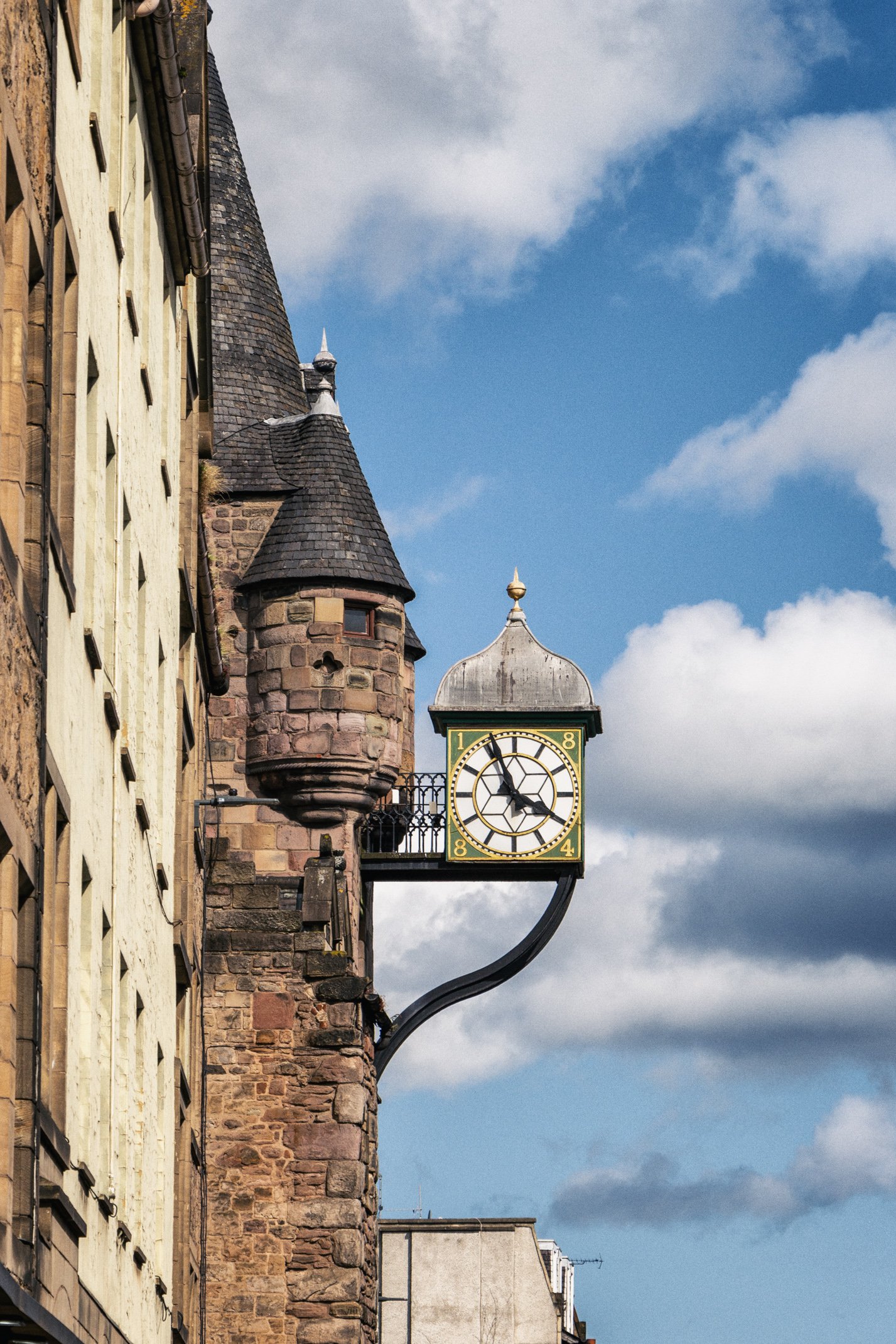 A historic building with a clock mounted on a black iron arm, featuring a round face with black Roman numerals and hands, against a partly cloudy blue sky.