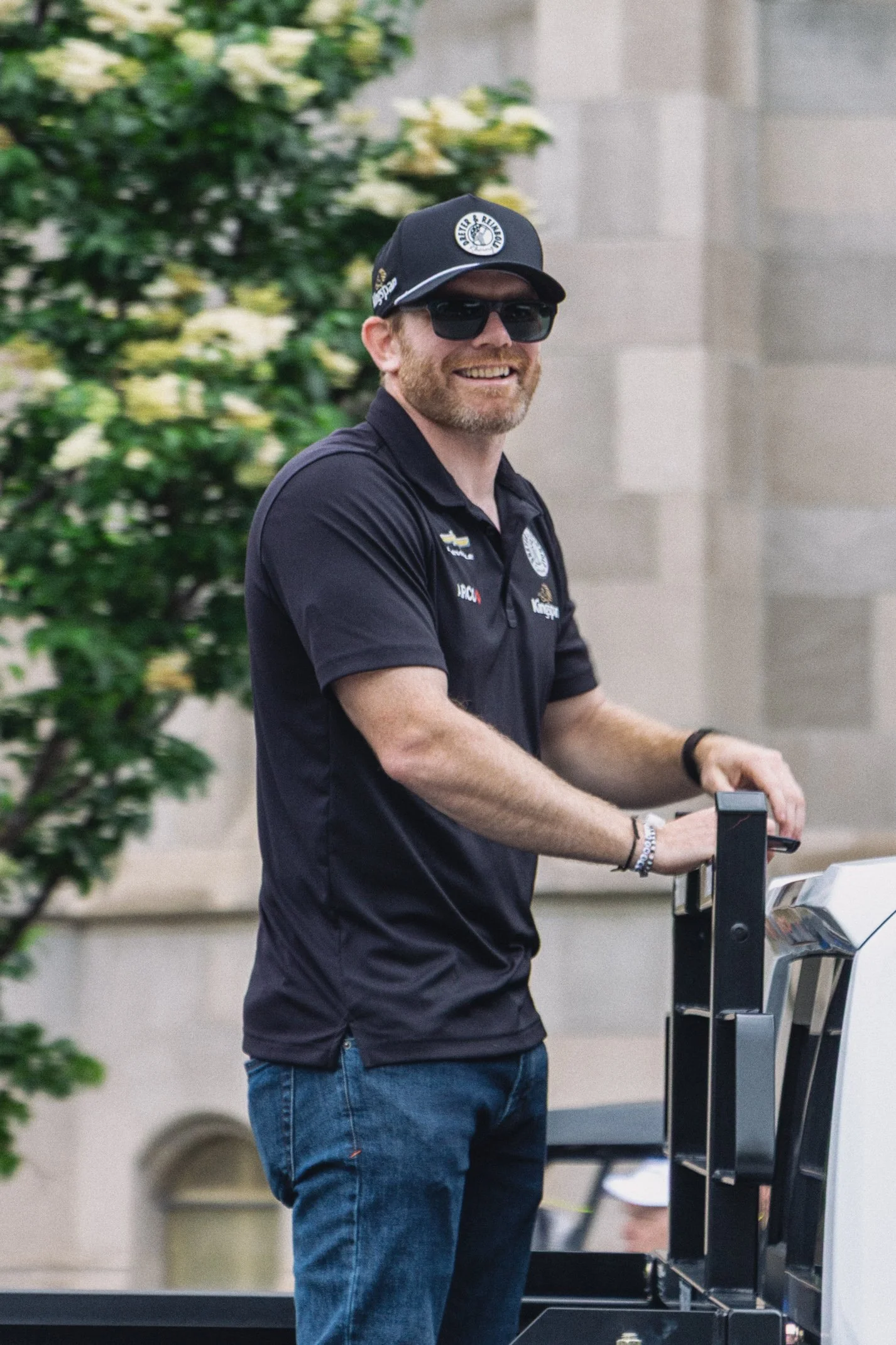 A man with sunglasses and a beard smiling while pushing a luggage cart outside, wearing a black polo shirt with logos, a black cap, and jeans, with green foliage and a stone wall in the background.