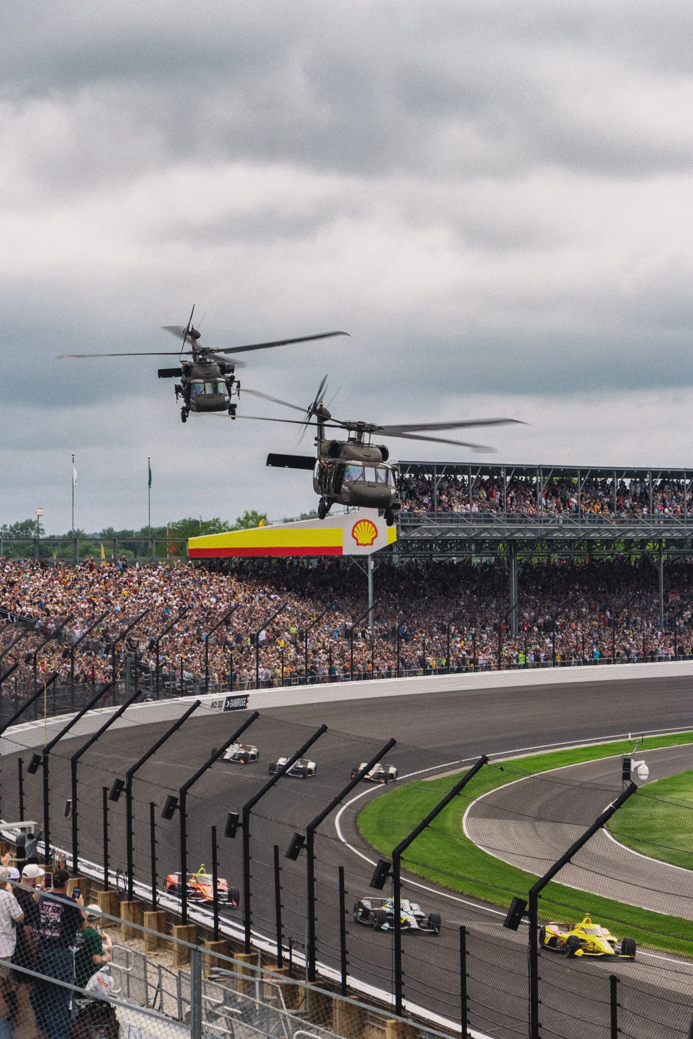 Two helicopters flying over a race track with cars racing below, crowd in the stands, and a cloudy sky.