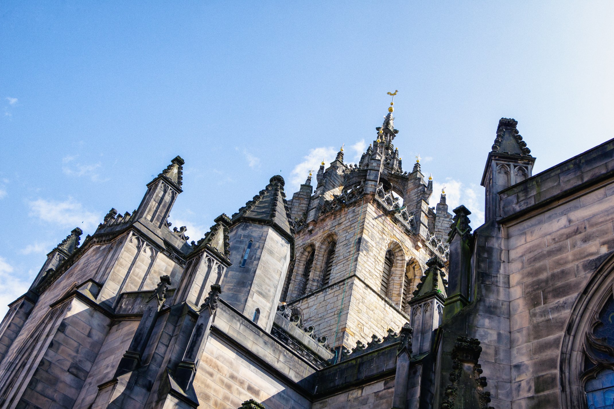 A close-up view of a historic Gothic-style cathedral's tower with ornate spires and a weather vane against a blue sky.