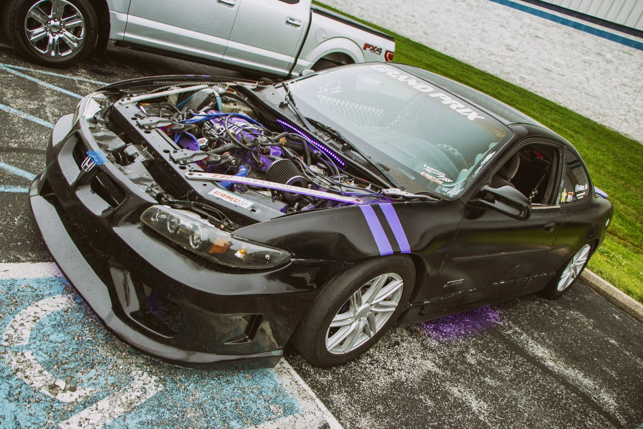 A black sports car with its hood open, revealing a purple and black engine, parked in a lot next to a silver pickup truck.