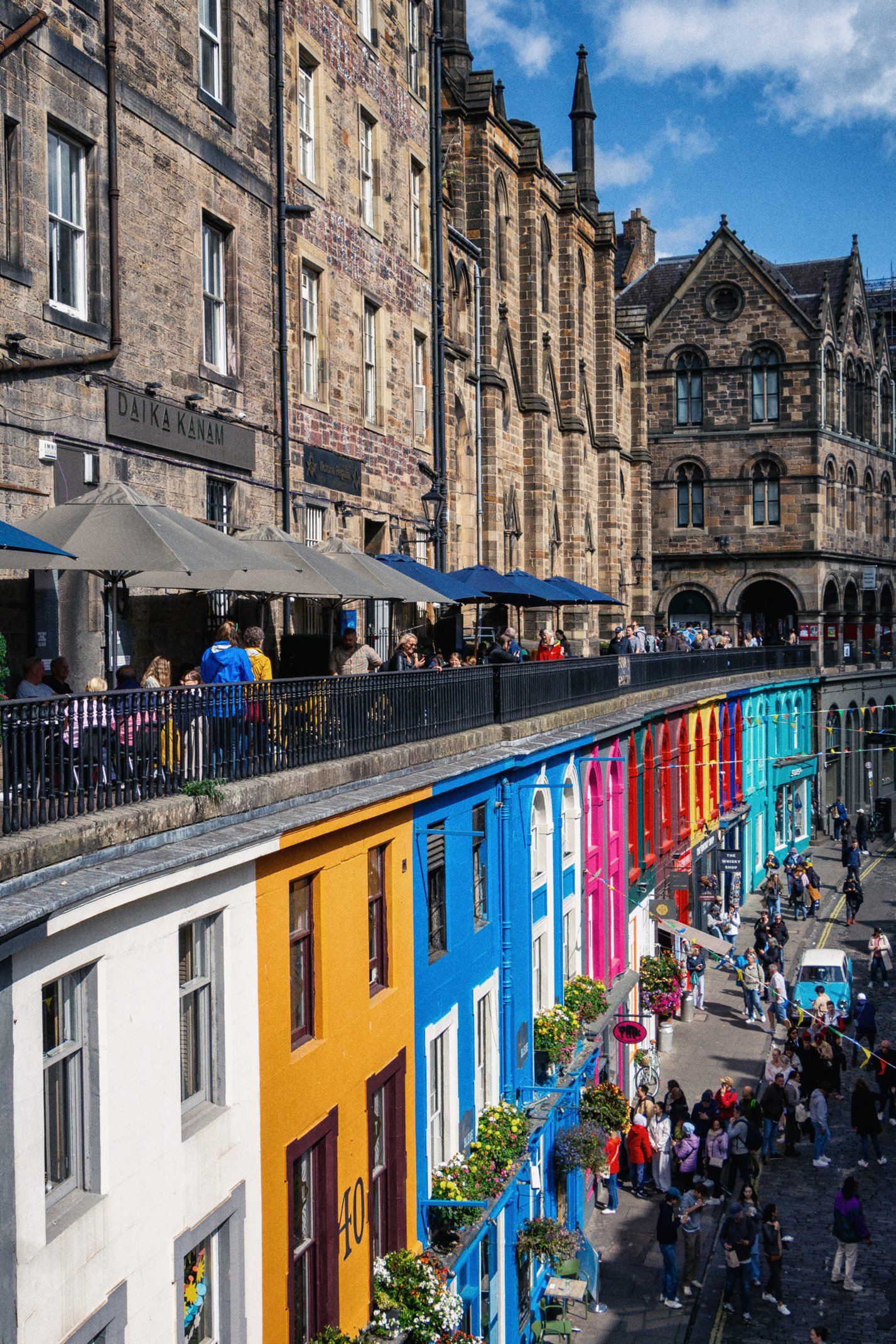 A busy street scene in Edinburgh, Scotland, with colorful buildings on the ground level, an outdoor cafe on the upper level, and people walking along the sidewalk.