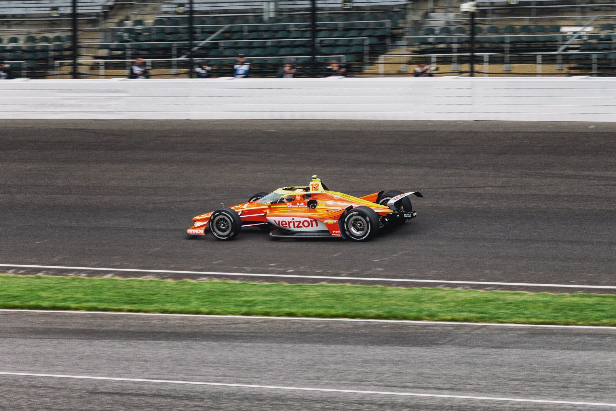 A racing car with Verizon branding on a race track, with spectators in the stands in the background.