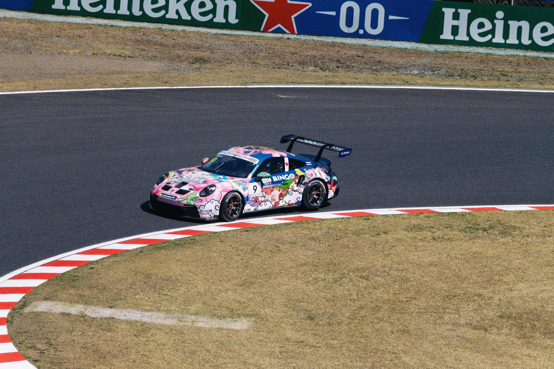 A race car on a track with a special colorful livery, featuring pink, purple, green, and white clouds and graphics, navigating a corner. There are Heineken and other advertisements in the background.