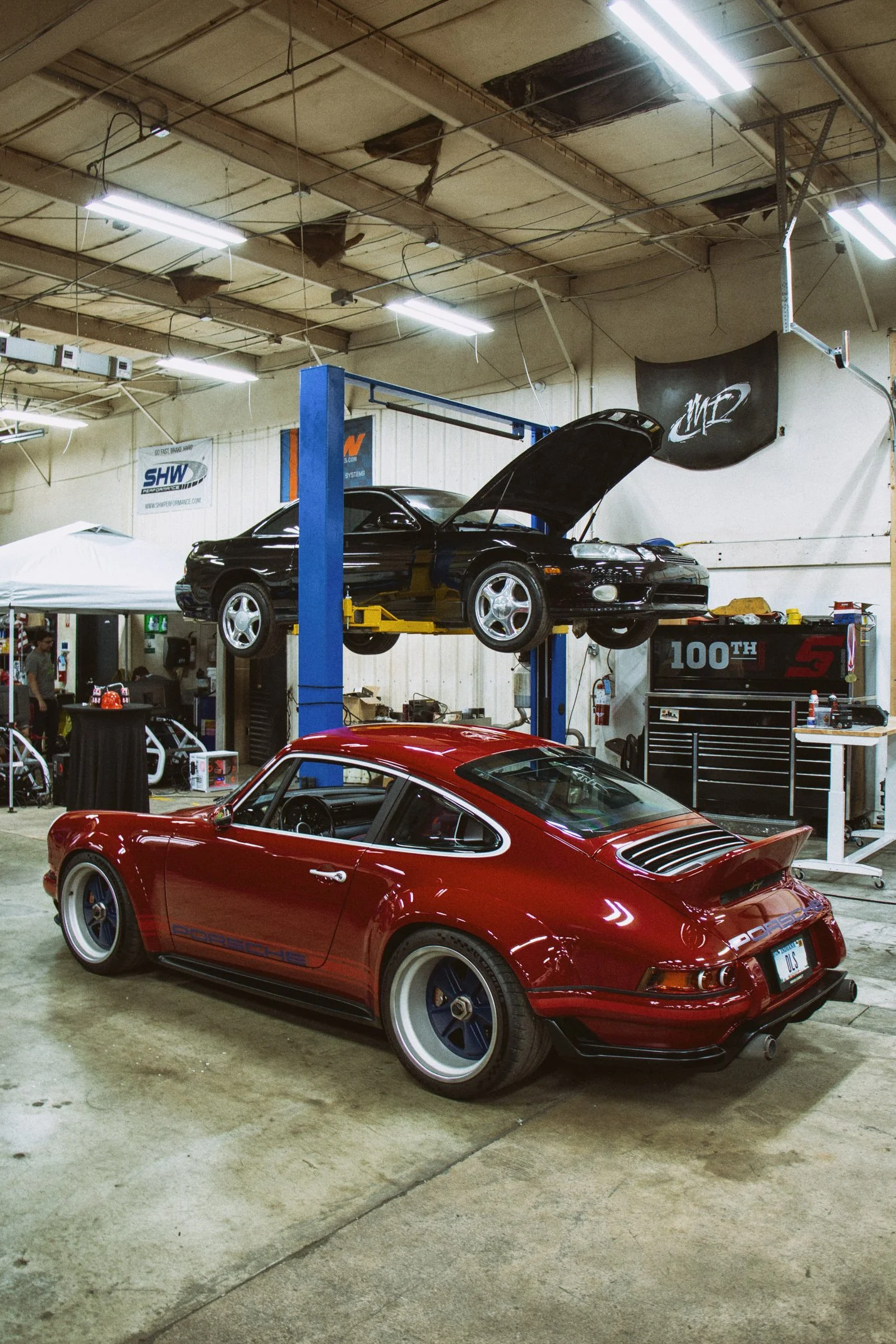 A red Porsche 911 sports car parked inside a workshop with another black car elevated on a hydraulic lift. The workshop has a damaged ceiling with torn insulation and fluorescent lights. There are various tools and equipment scattered around.