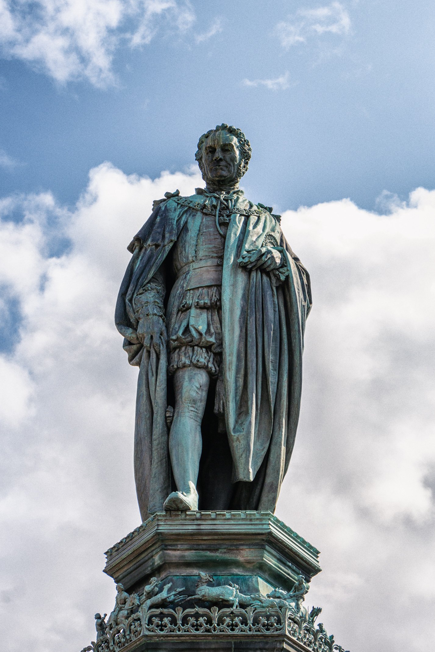A statue of a historical figure dressed in regal clothing, standing on a decorative pedestal against a blue sky with clouds.