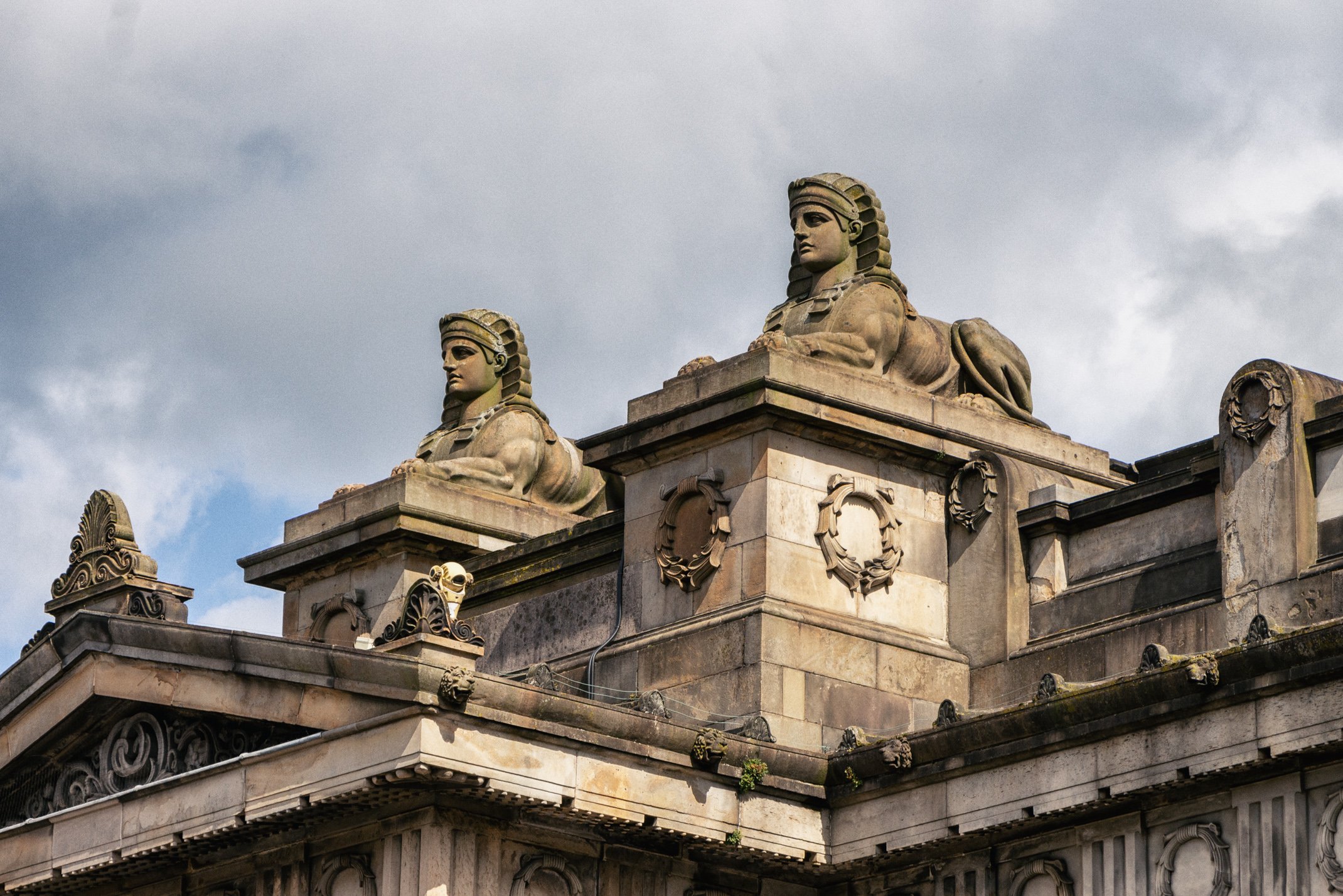 Close-up of stone sculptures of sphinxes on the rooftop of a historic building with overcast sky in the background.