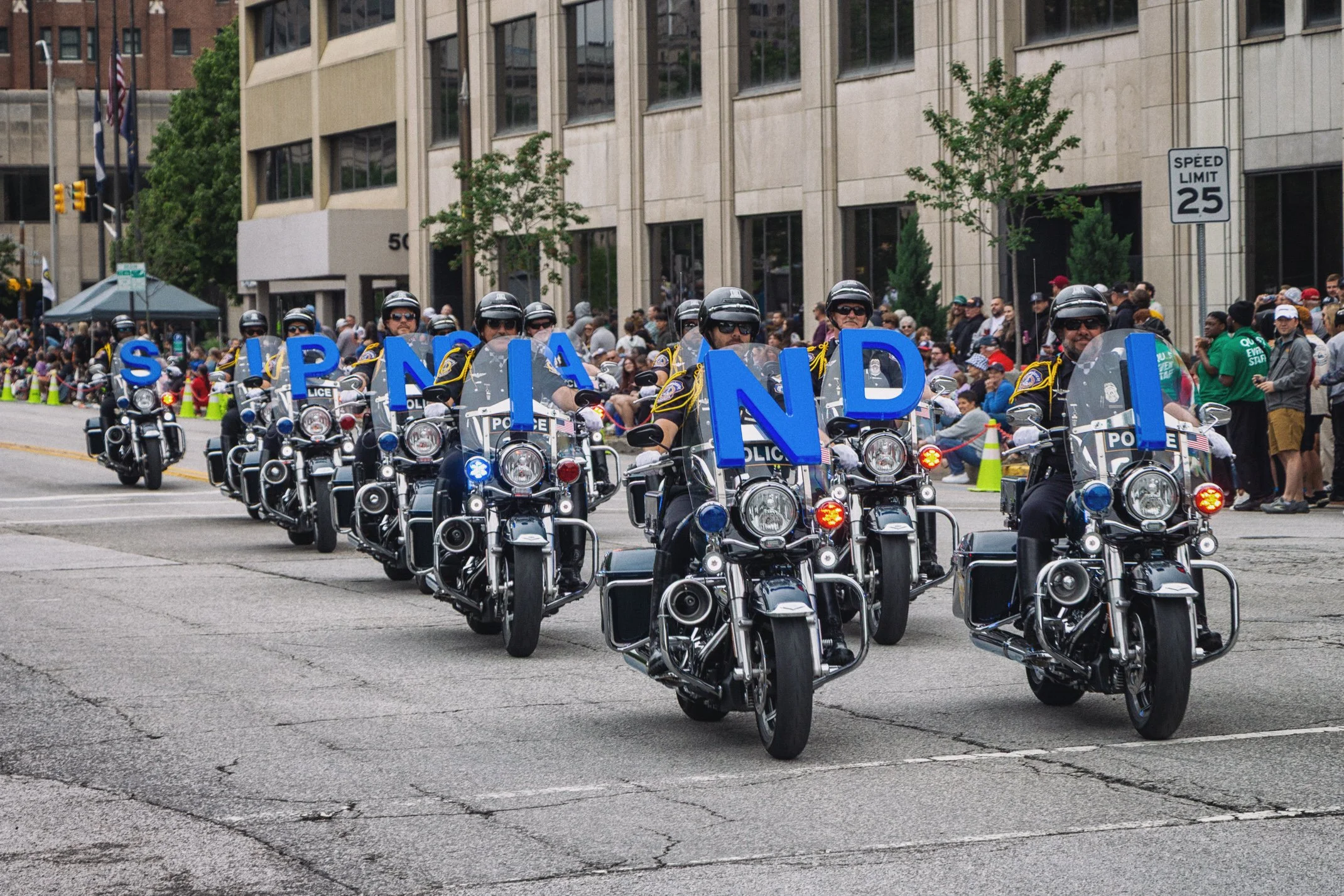 A line of police officers on motorcycles forming a parade or procession on a city street, with a crowd of spectators and tall buildings in the background.