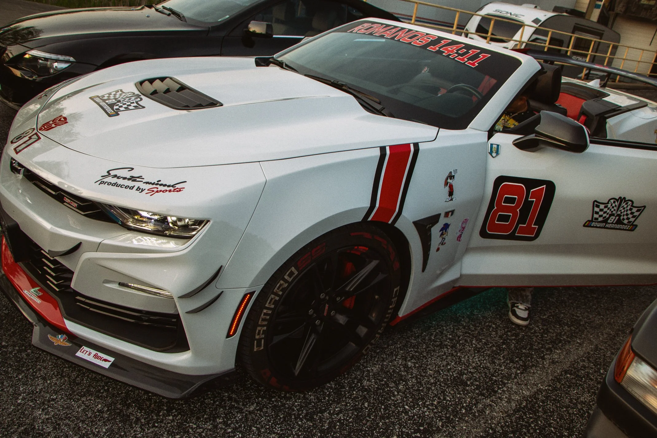 White race car with racing decals, black accents, a large rear wing, parked beside a black car and a person wearing sneakers.