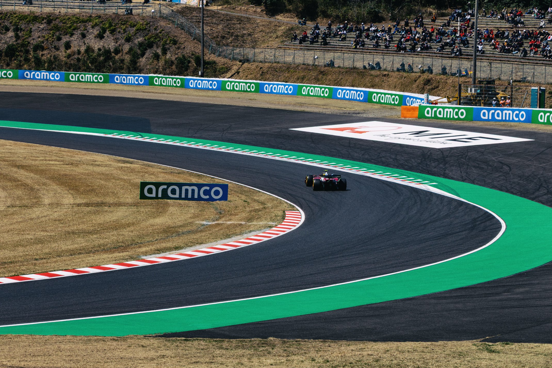 A single race car navigating a curving section of a race track with green and white painted curbs, surrounded by desert terrain and stands filled with spectators. Advertising banners line the track.