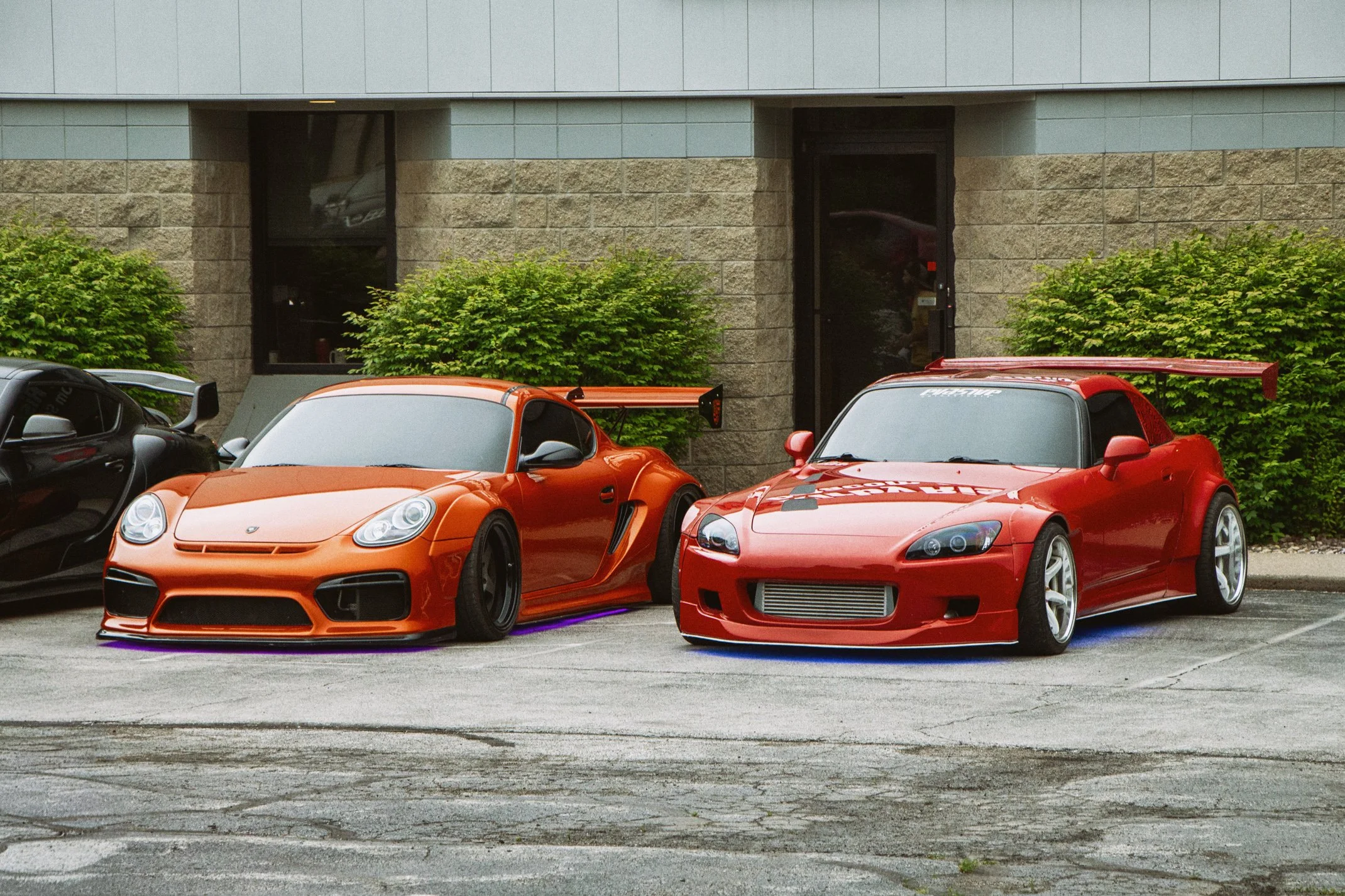 Three sports cars parked on the street in front of a building with bushes. The middle car is orange, the right car is red, and part of a black car is visible on the left.