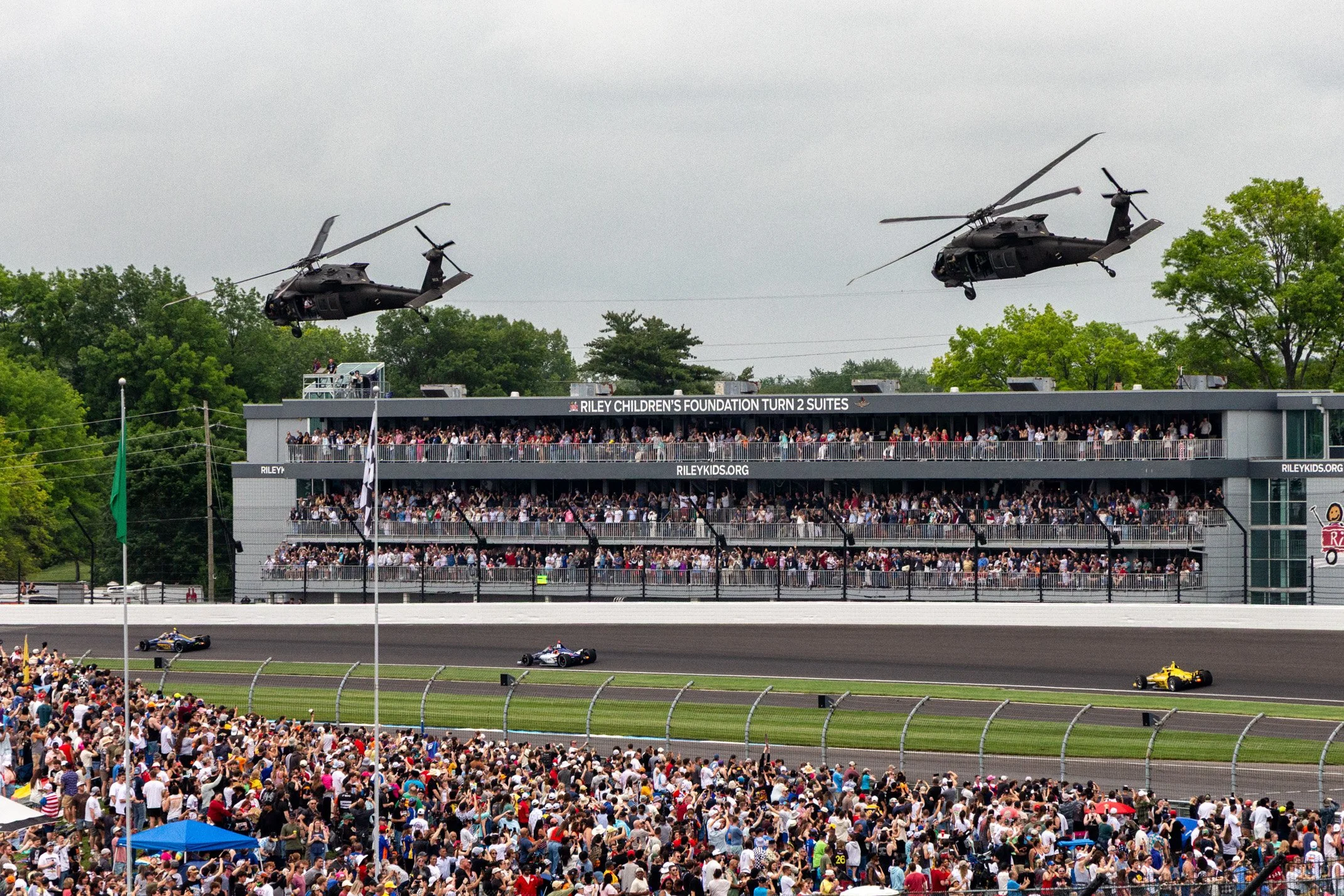 Race track with a large crowd watching cars race, two helicopters flying overhead, and a multi-story building with spectators.