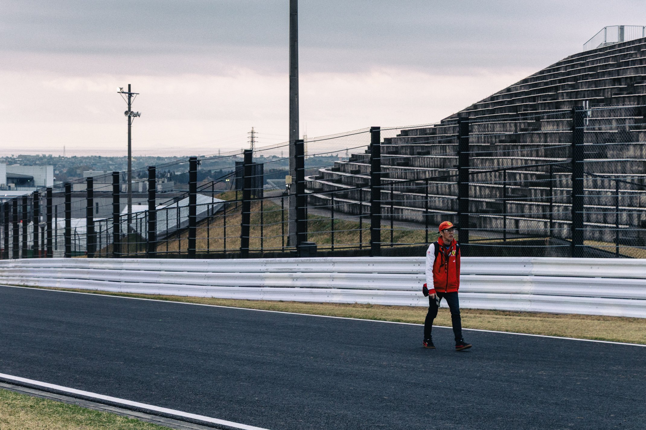 A man walking on a race track wearing a red jacket, black pants, and a red cap, with grandstands and fencing in the background under a cloudy sky.