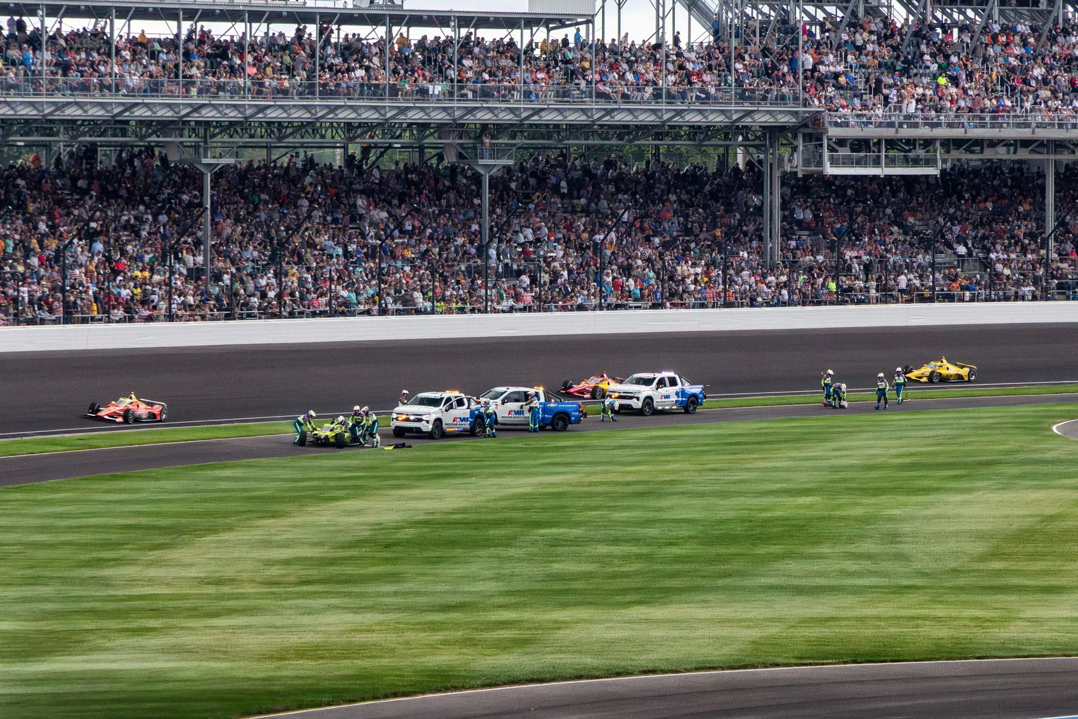 Race track with a yellow race car and safety vehicles including police and emergency vehicles, and several personnel attending to an incident, with packed grandstands of spectators in the background.