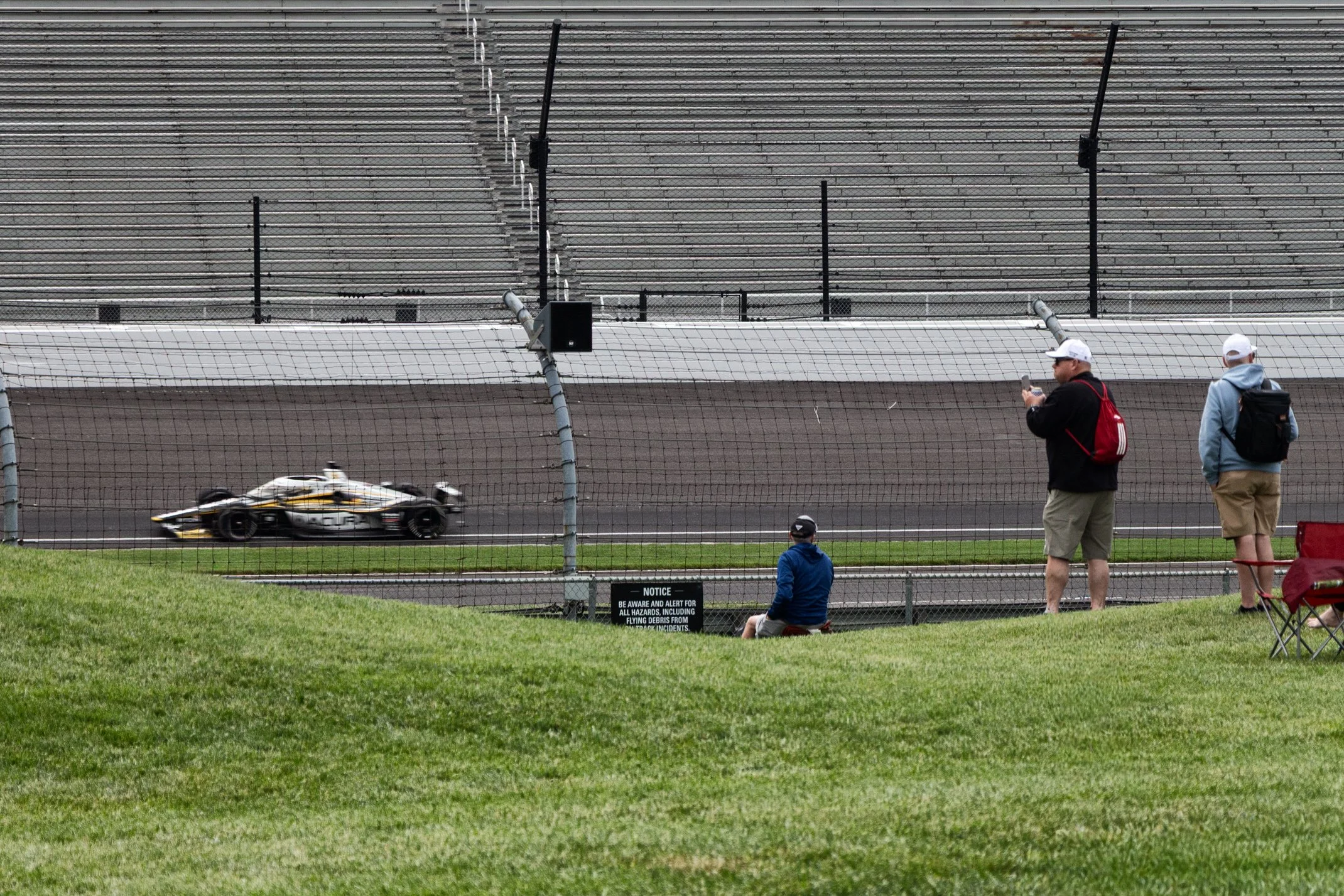 A race car on a track with people watching behind a fence, some taking photos and others seated on the grass, with empty bleachers in the background.