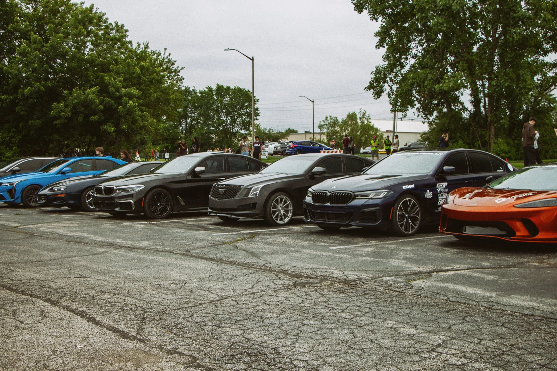 A parking lot with various luxury cars parked in front of a group of people standing and walking, some wearing safety vests, with trees and overcast sky in the background.